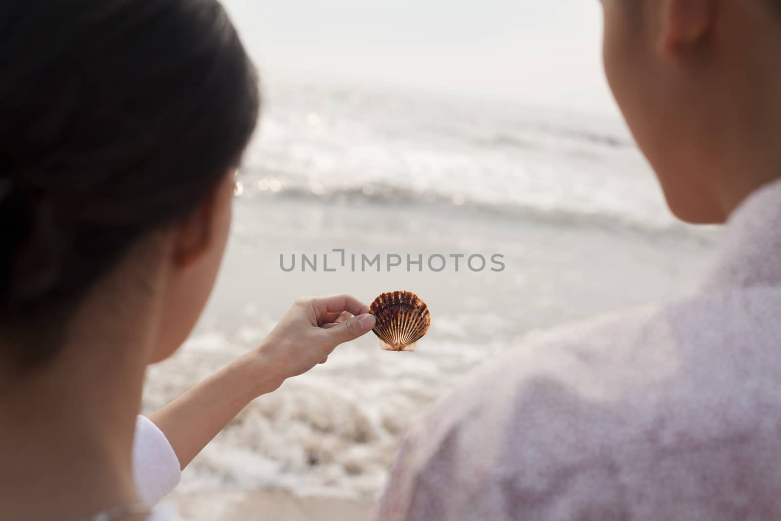 Young couple standing and looking at seashell on the beach, over the shoulder view