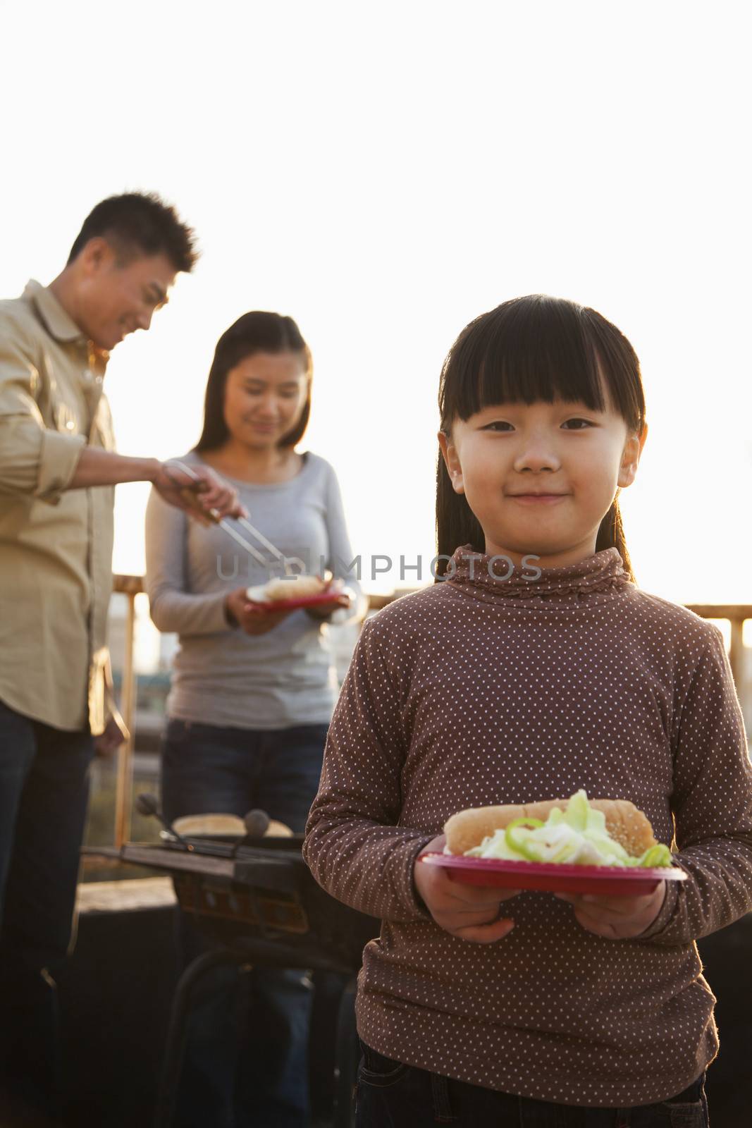 daughter holding plate with hotdog and salad, parents standing next to the barbeque by XiXinXing