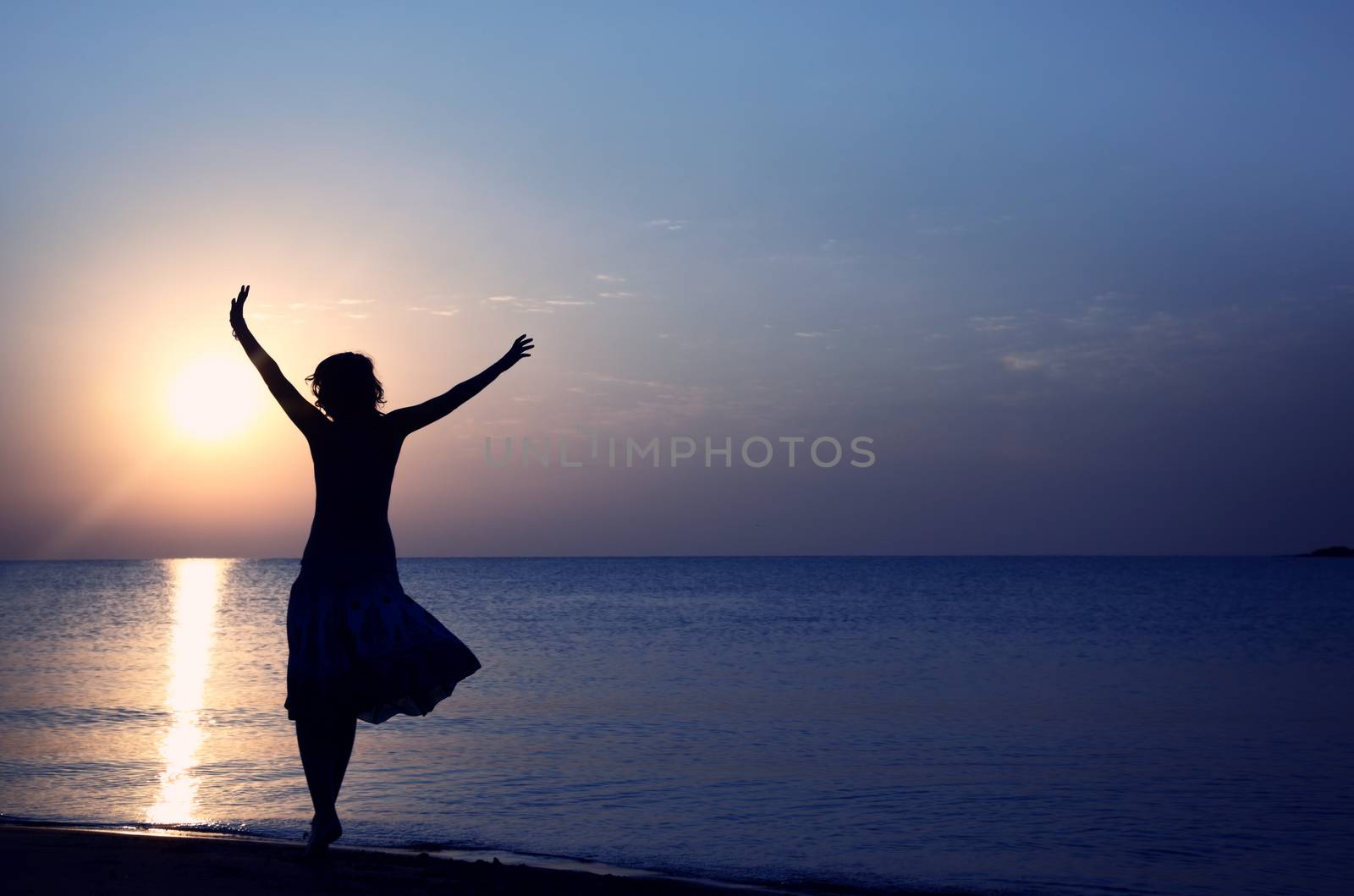 Silhouette of the happy woman in summer dress standing at the beach during sunset. Natural light and colors