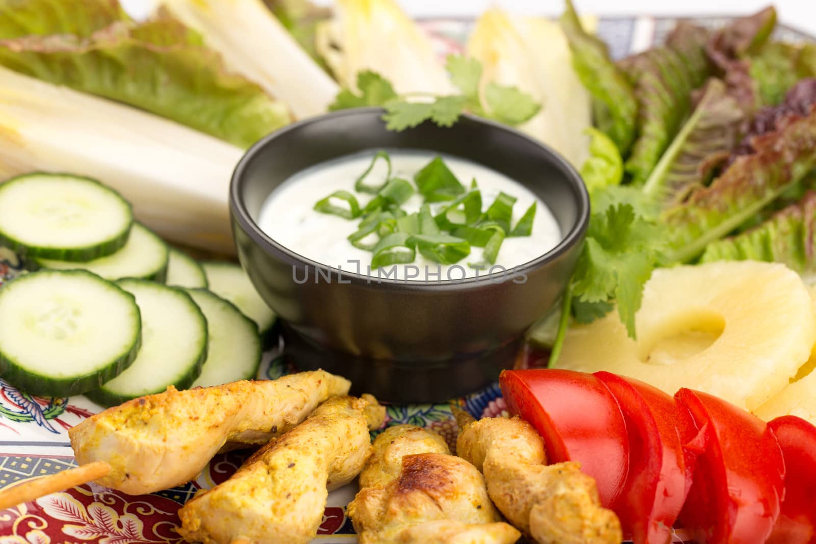 Asian food with chicken skewers, sauce, tomatoes, cucumber, pineapples, endives and salad.