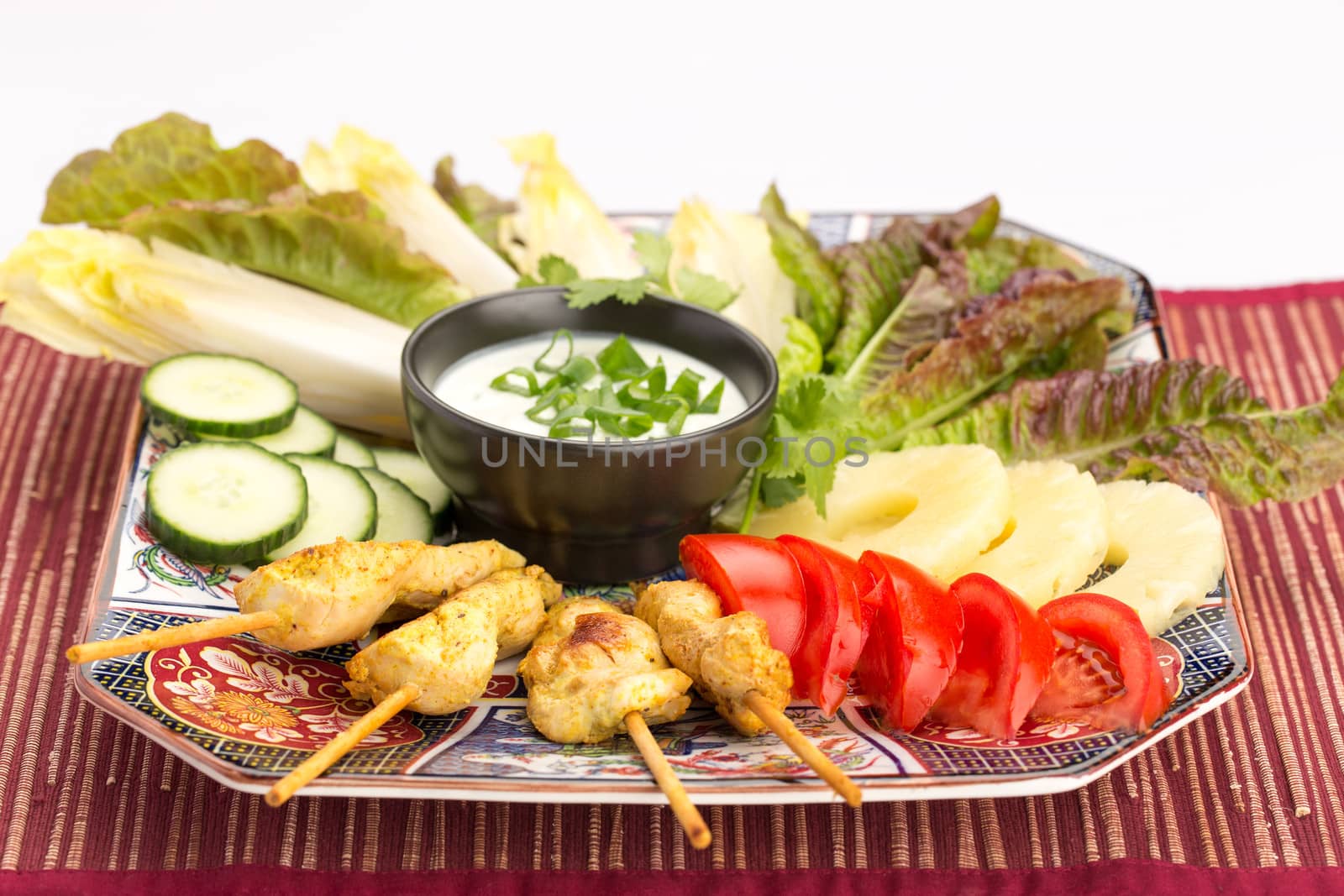 Asian food with chicken skewers, sauce, tomatoes, cucumber, pineapples, endives and salad.