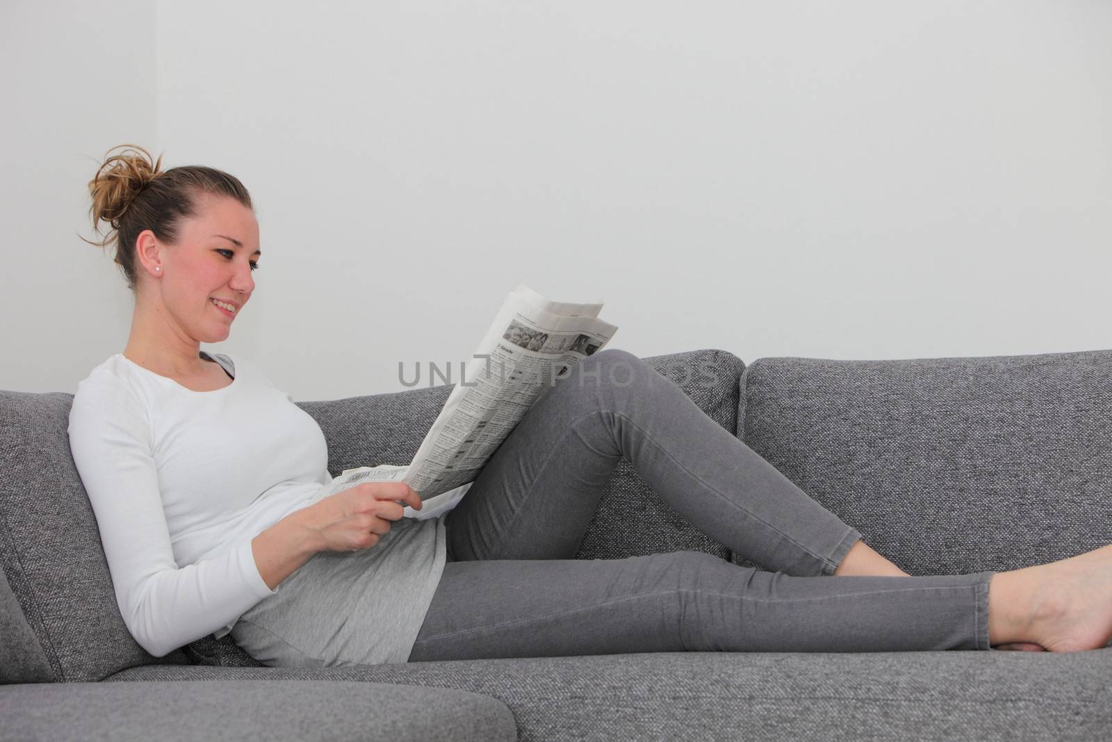 Pretty young woman relaxing on a sofa at home reading a newspaper with copyspace