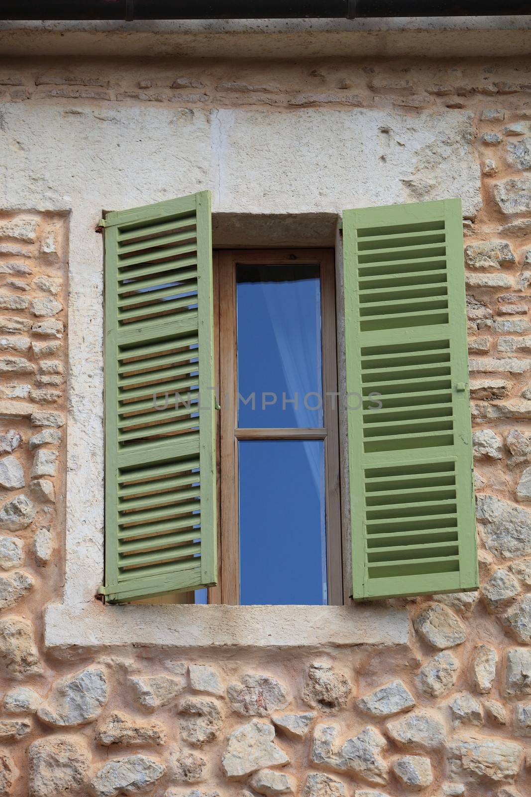 Window with green wooden shutters half open in a stone building with walls made of natural rocks