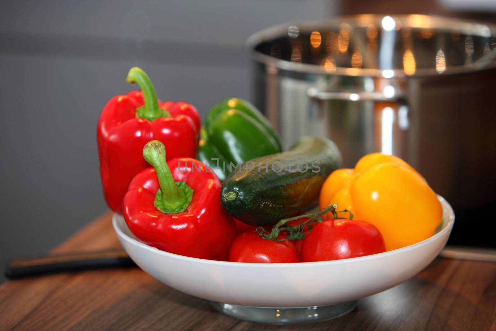 Brightly coloured bowl of vegetables with red, green and yellow bell peppers and tomatoes standing on a kitchen table near the stove and a saucepan