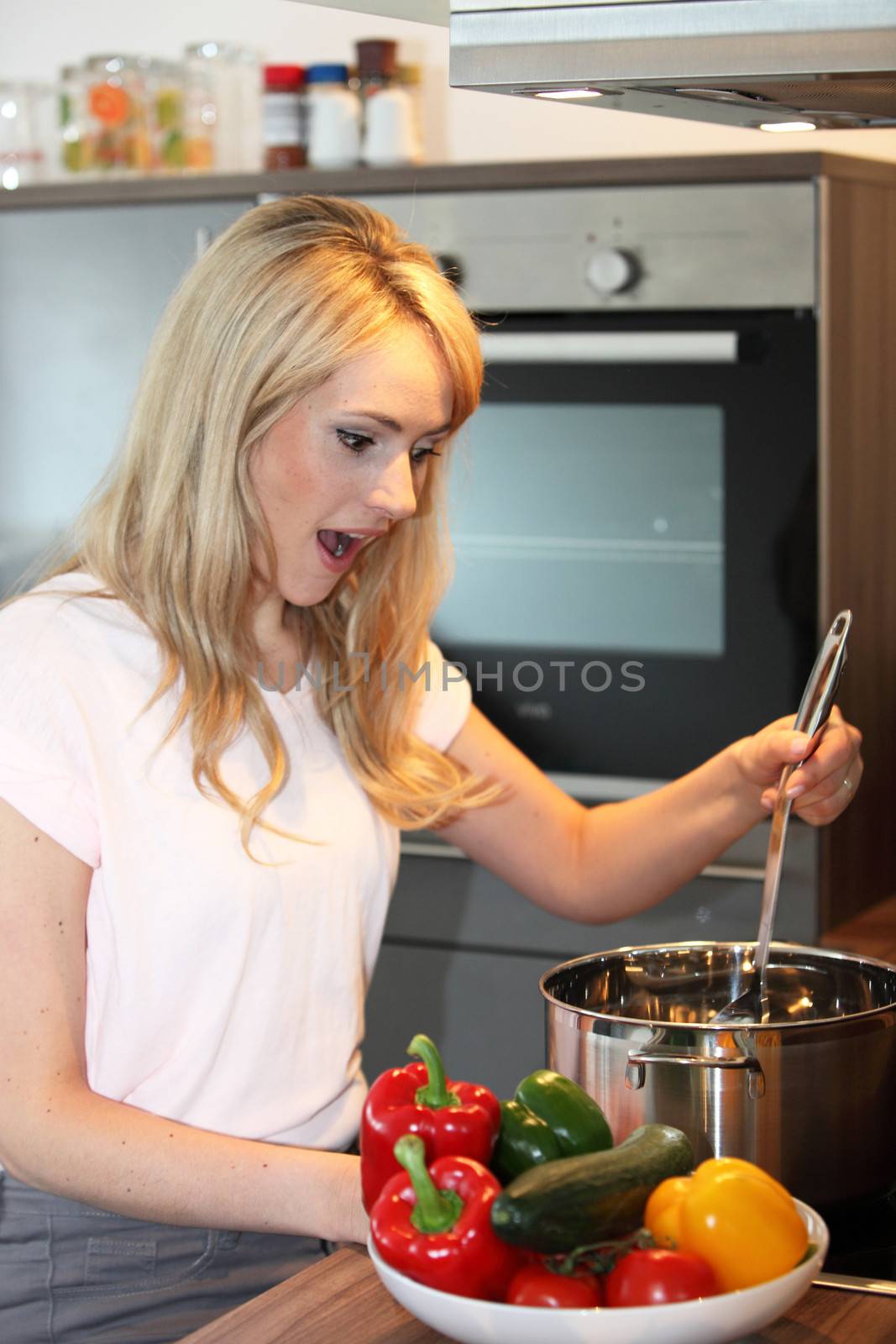 Attractive young blond woman cooking in the kitchen stirring the pot over the stove with a surprised look and a colourful bowl of fresh vegetables in the foreground