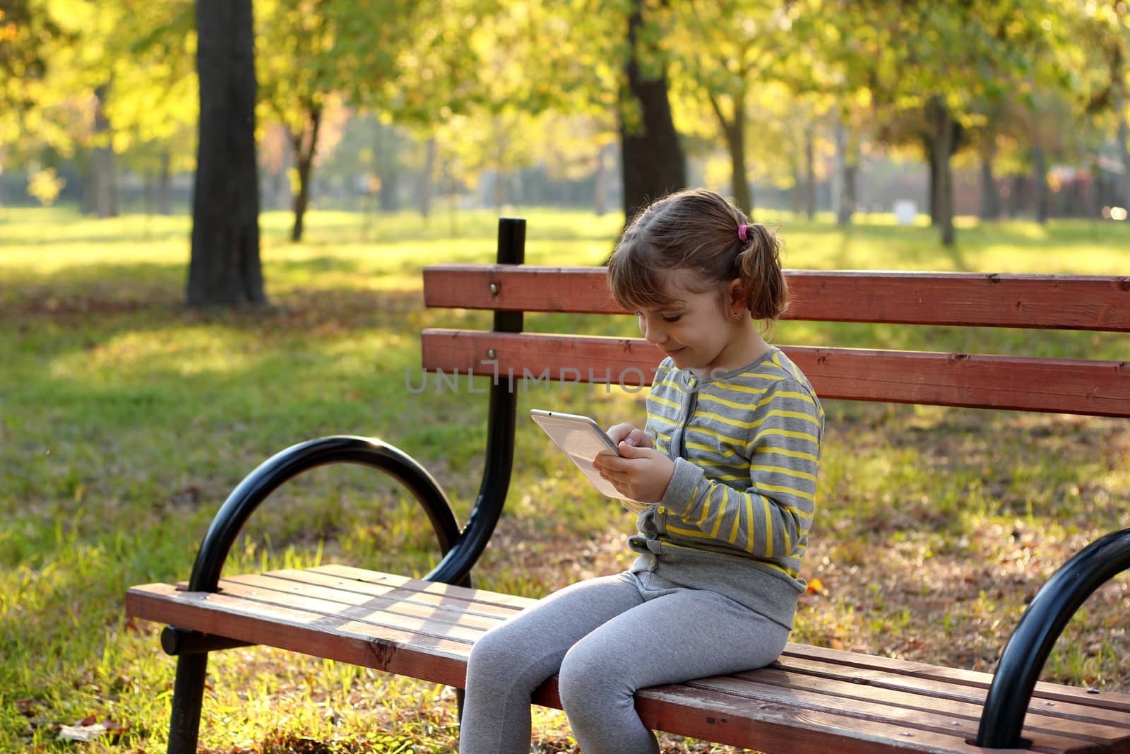 little girl play with tablet in autumn park by goce