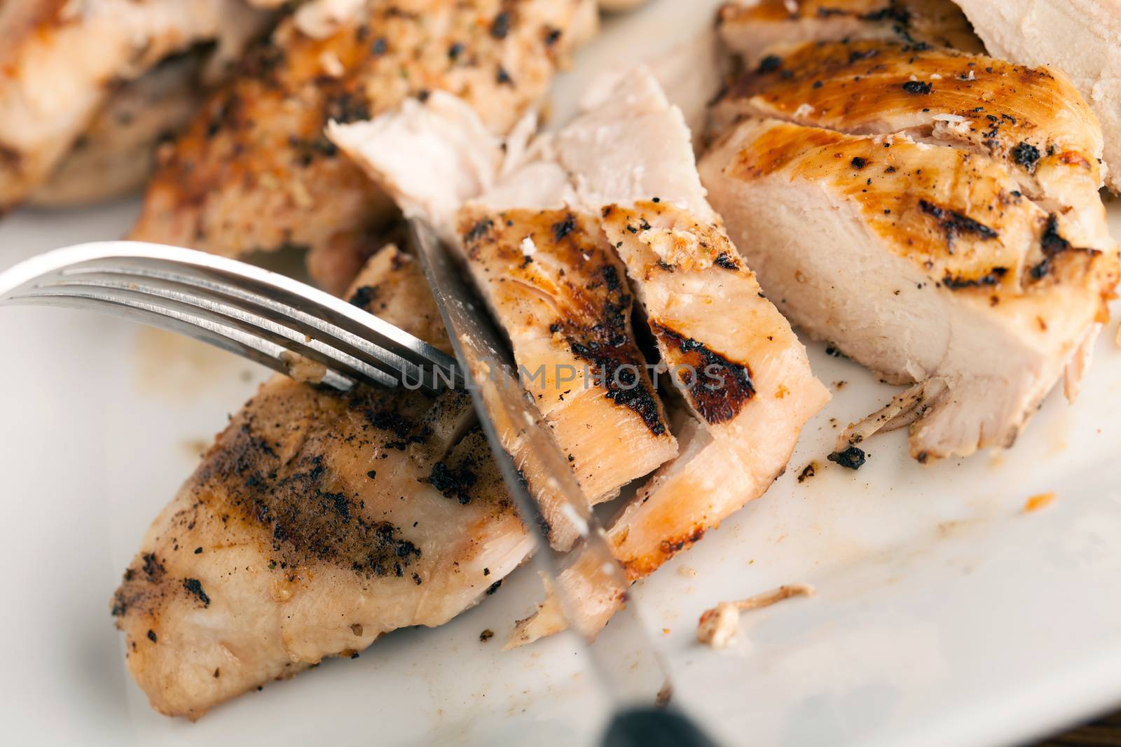 Freshly prepared grilled chicken being cut up with a fork and knife. First person point of view. Shallow depth of field.