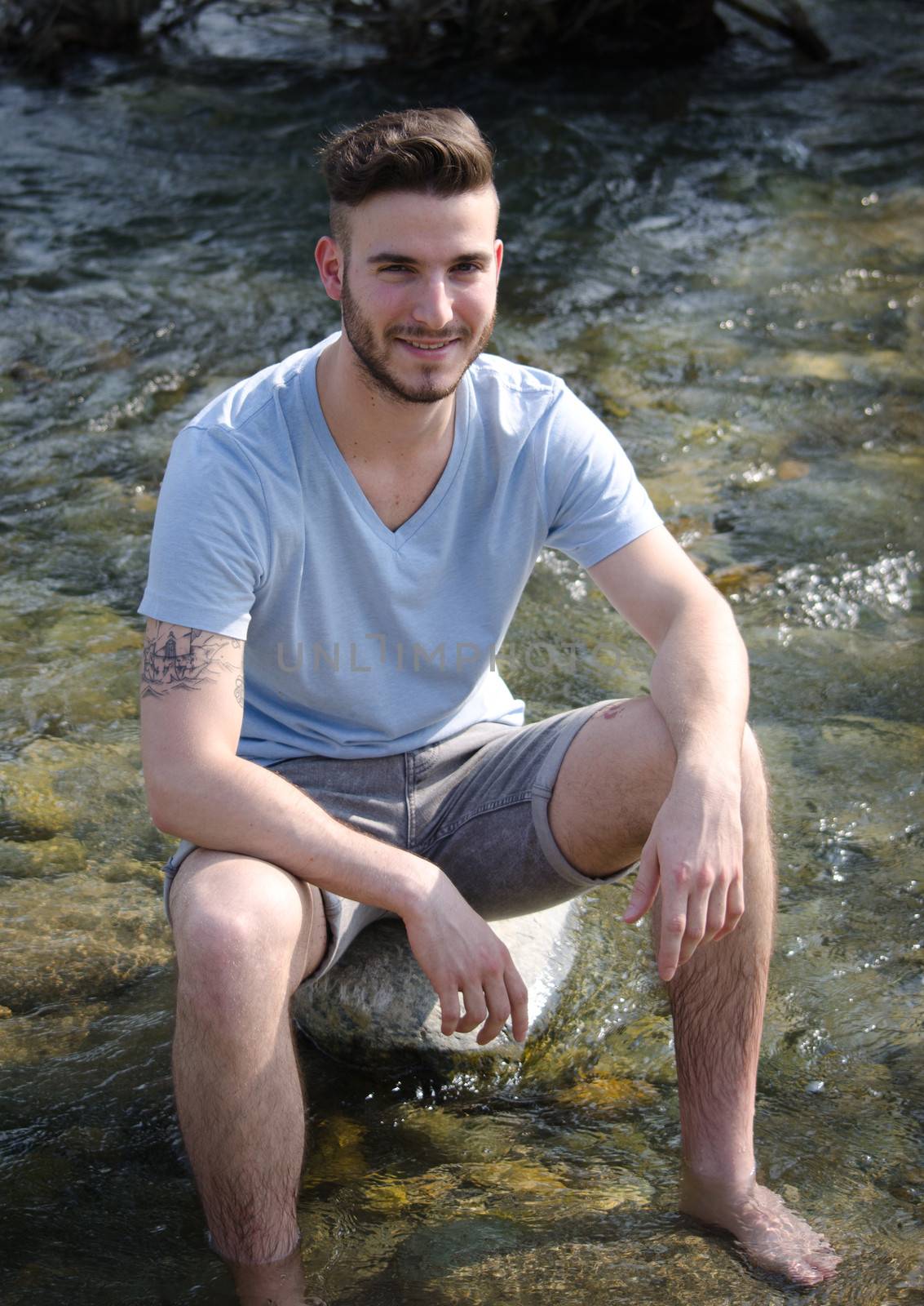 Attractive young man sitting on rock in a river, smiling with feet in the water