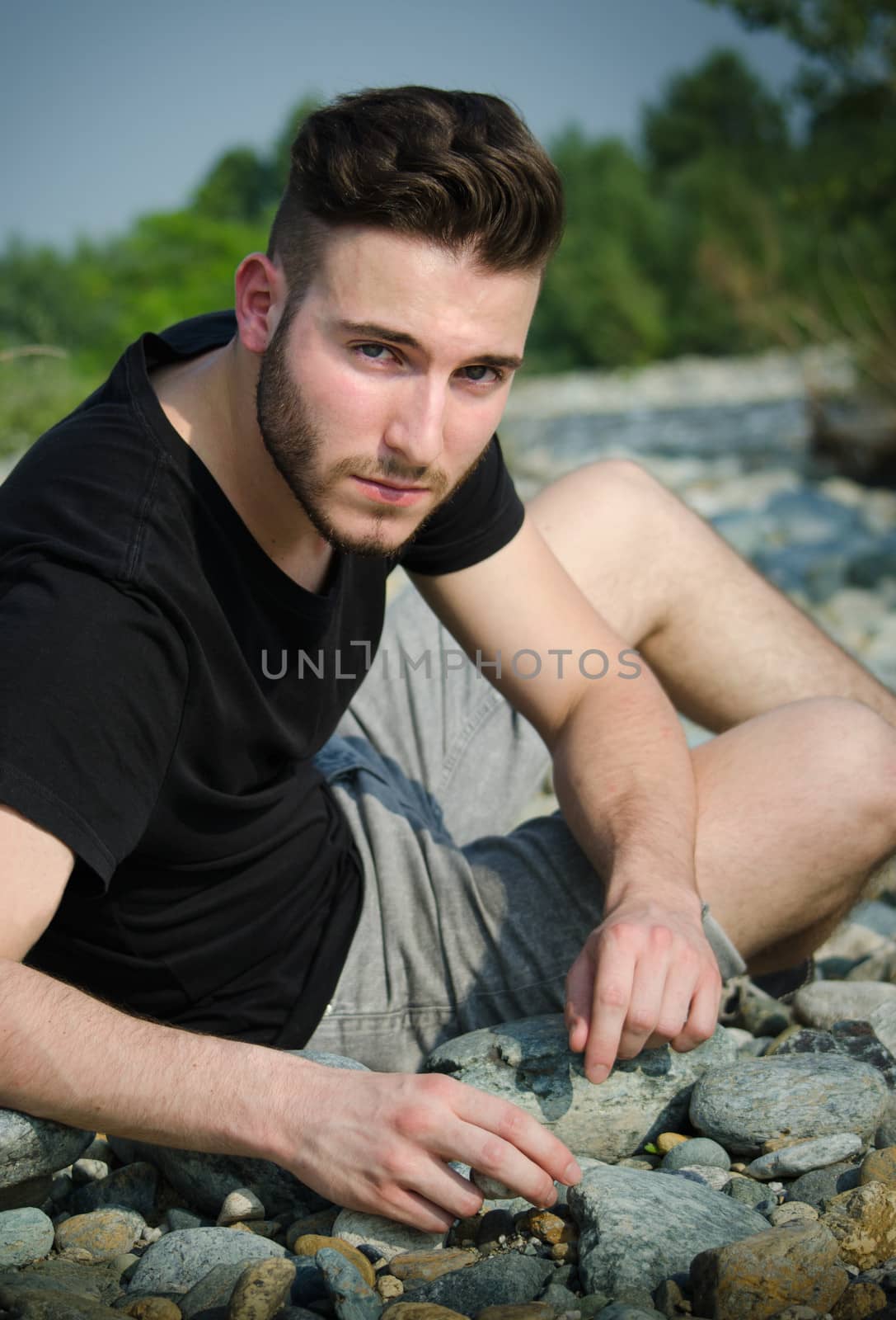 Handsome young man outdoors, sitting on rock, stones and pebbles