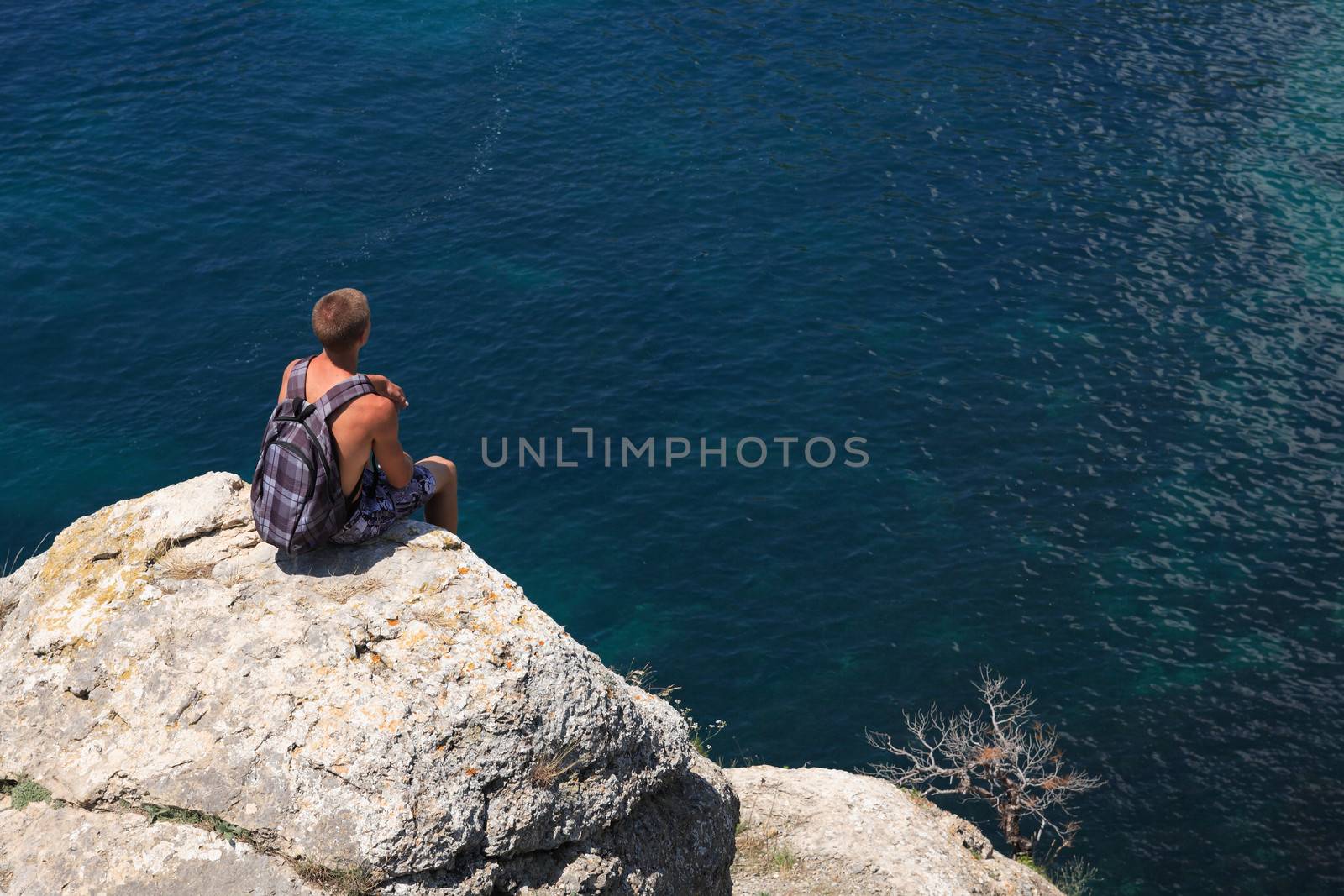 Lonely young hiker sitting on top of the rock and looking to the blue sea