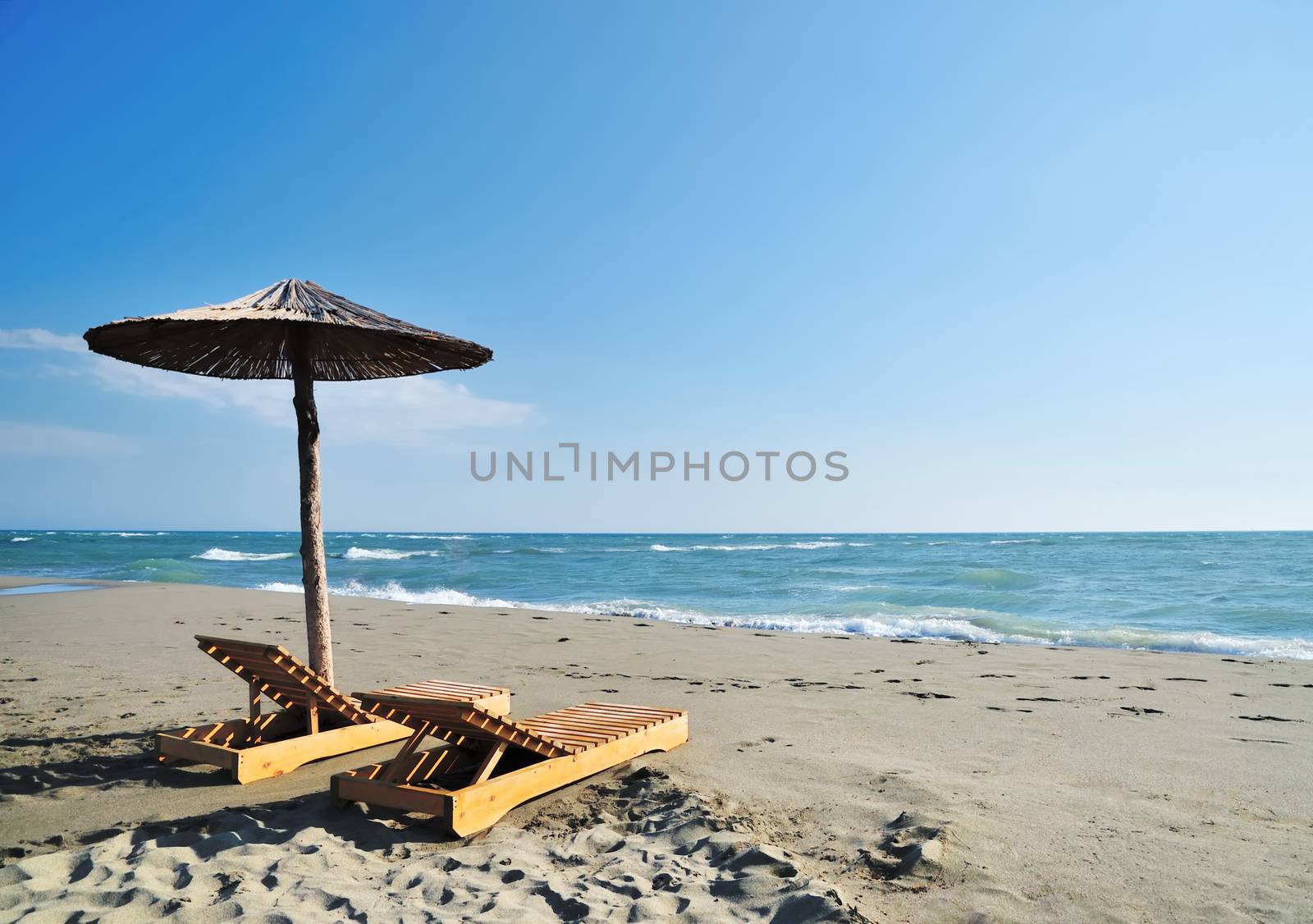 Two wooden deckchairs under straw parasol on sandy beach