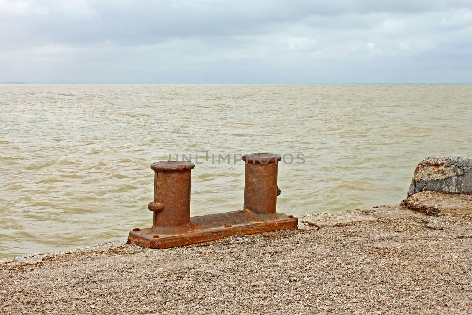 Steel rusty dual berth bitt on a fragment of old concrete pier on the background yellowish sandy seawater after the st