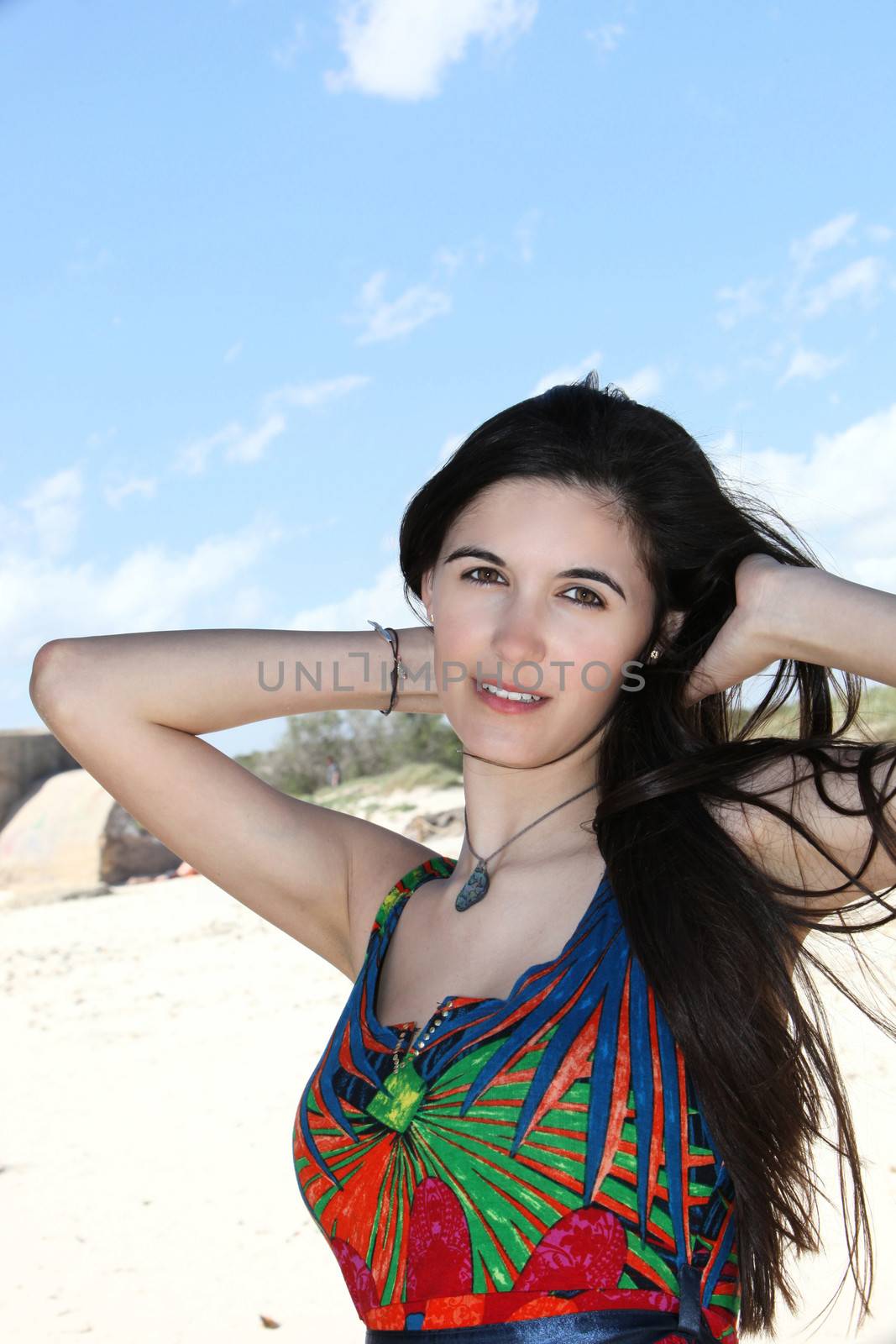 Beautiful woman on a summer vacation at the beach posing with her hands raised behind her neck in her long straight brunette hair wearing a colourful summer dress