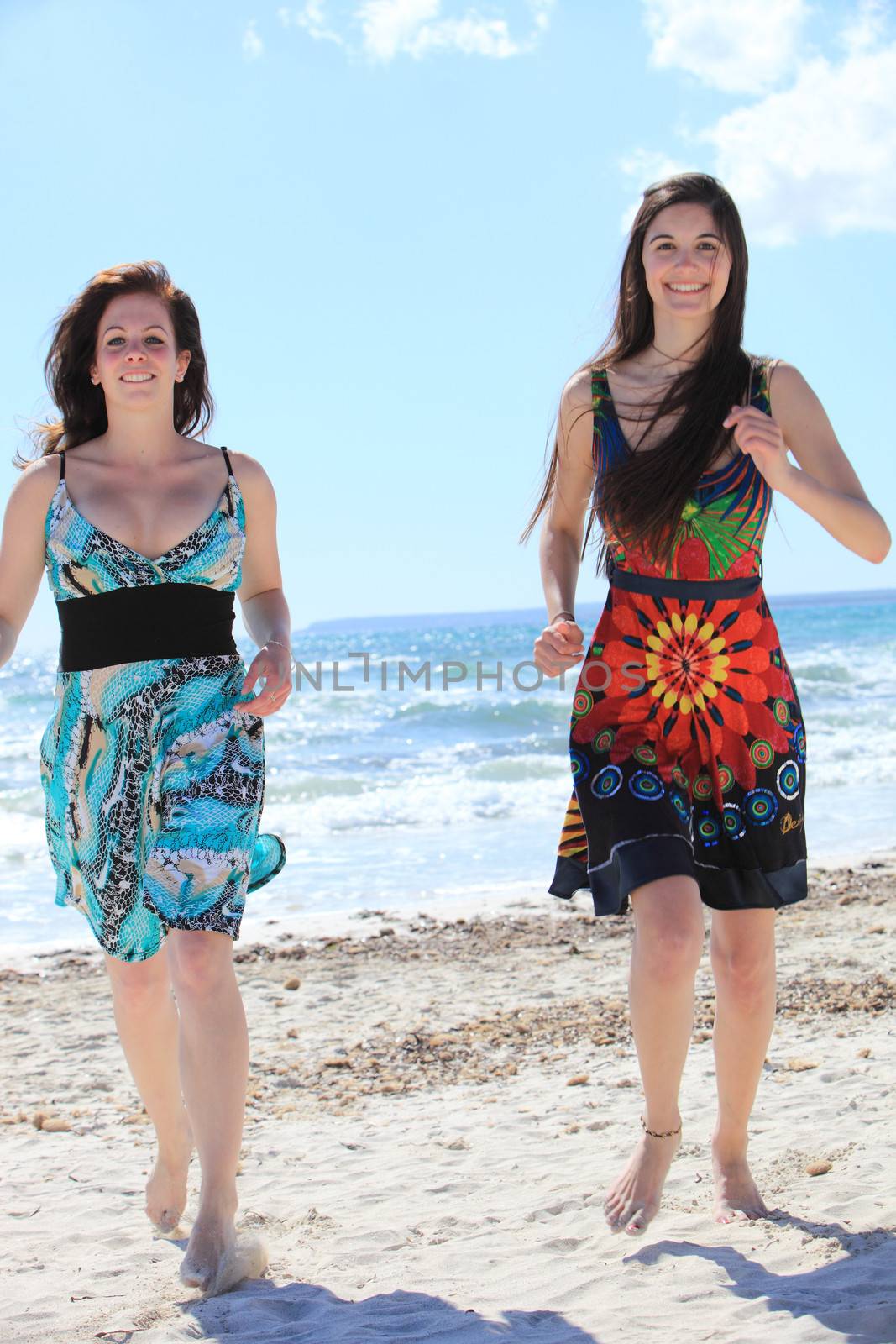 Two attractive barefoot women on the beach in their colourful summer dresses walking fast towards the camera across the sand with the sea behind them