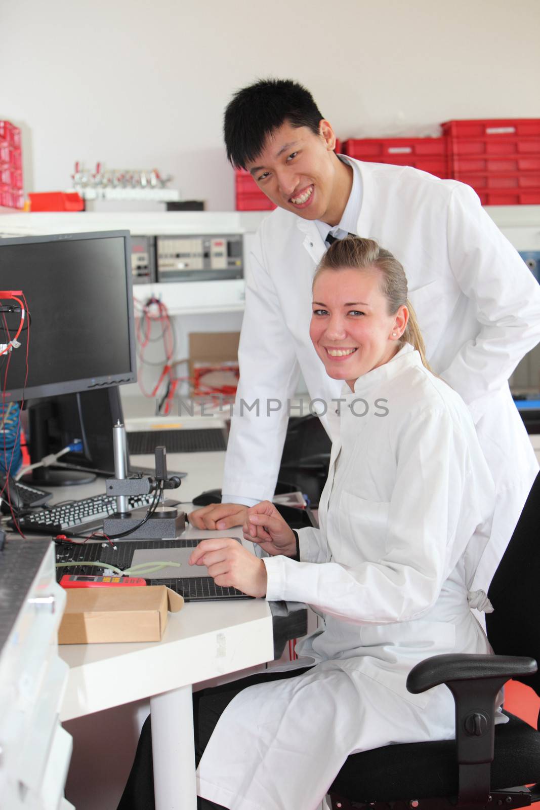 Two technicians at work in a laboratory with a young Asian man standing over his attractive female colleague as they discuss a test result