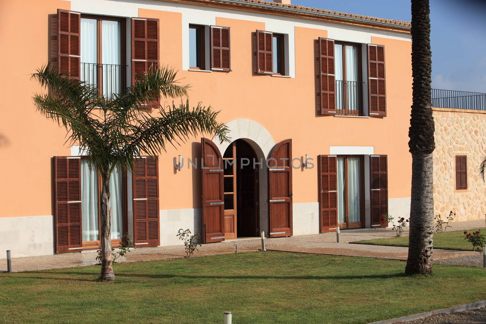 Entrance to an upmarket villa with a decorative painted facade and wooden shutters on an arched doorway and tall windows with a palm tree in the garden