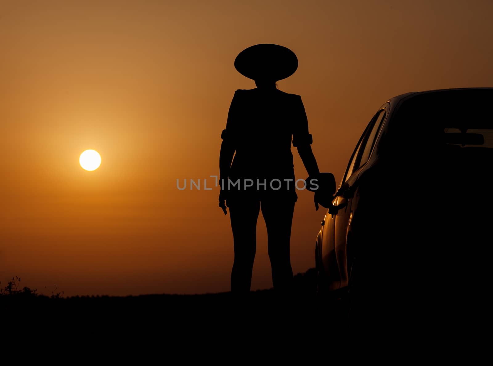 Silhouette woman with hat standing near car, against orange sunset