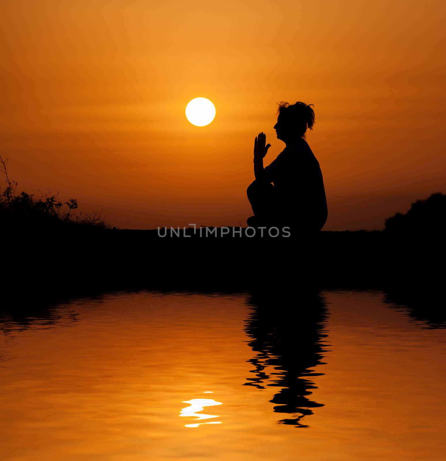 Silhouette woman sitting and relaxing against orange sunset with reflection in water