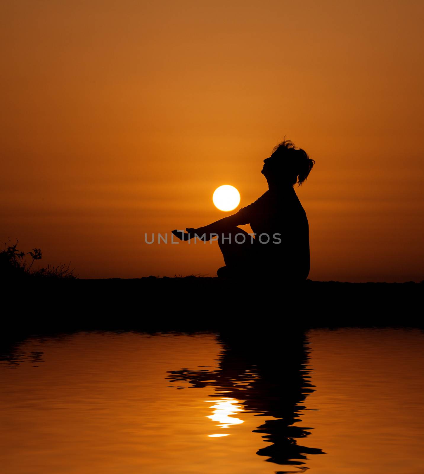 Silhouette woman sitting and relaxing against orange sunset with reflection in water