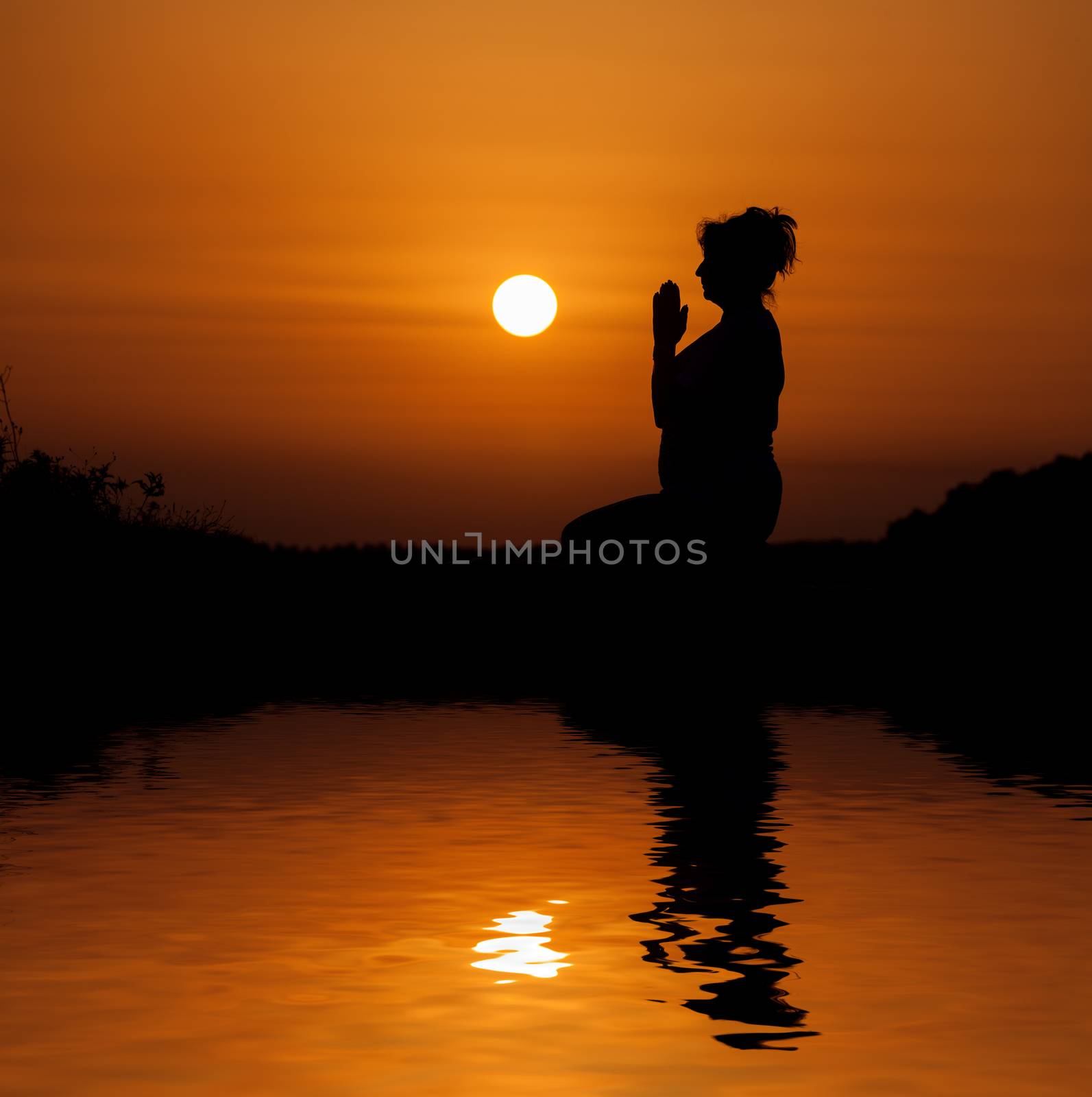 Silhouette woman sitting and relaxing against orange sunset with reflection in water