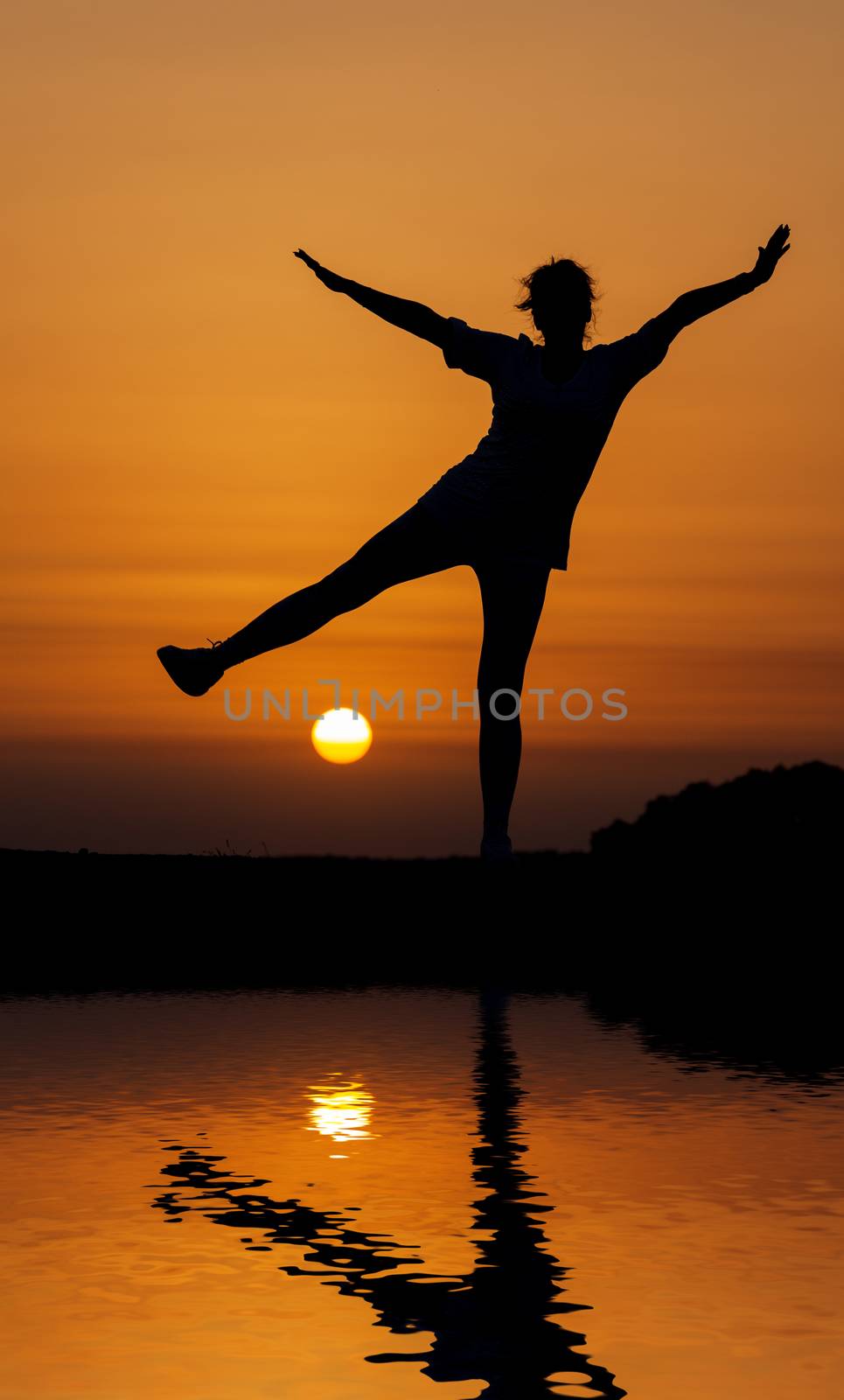 Silhouette woman jumping against orange sunset with reflection in water