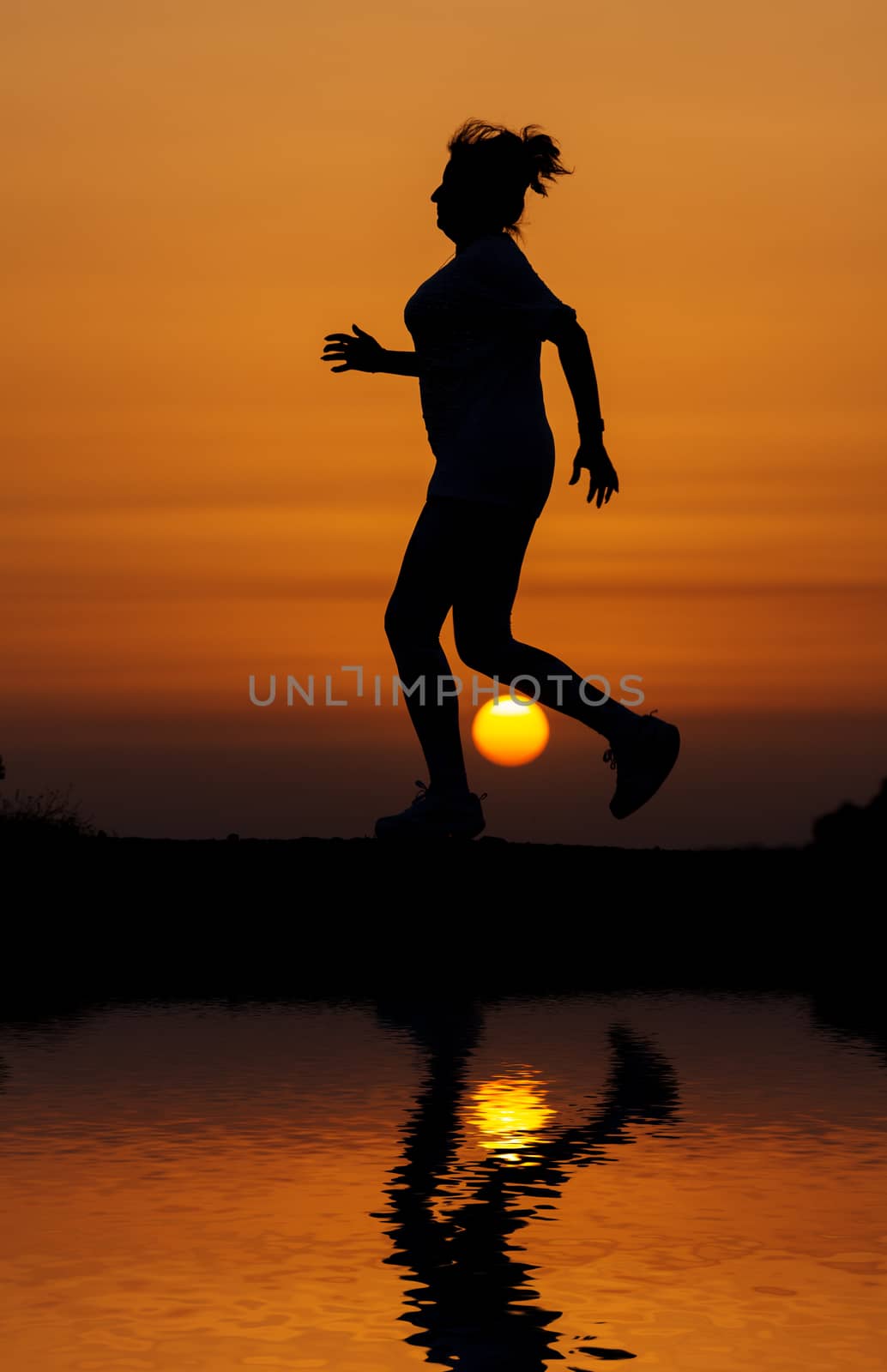 Silhouette woman running against orange sunset with reflection in water