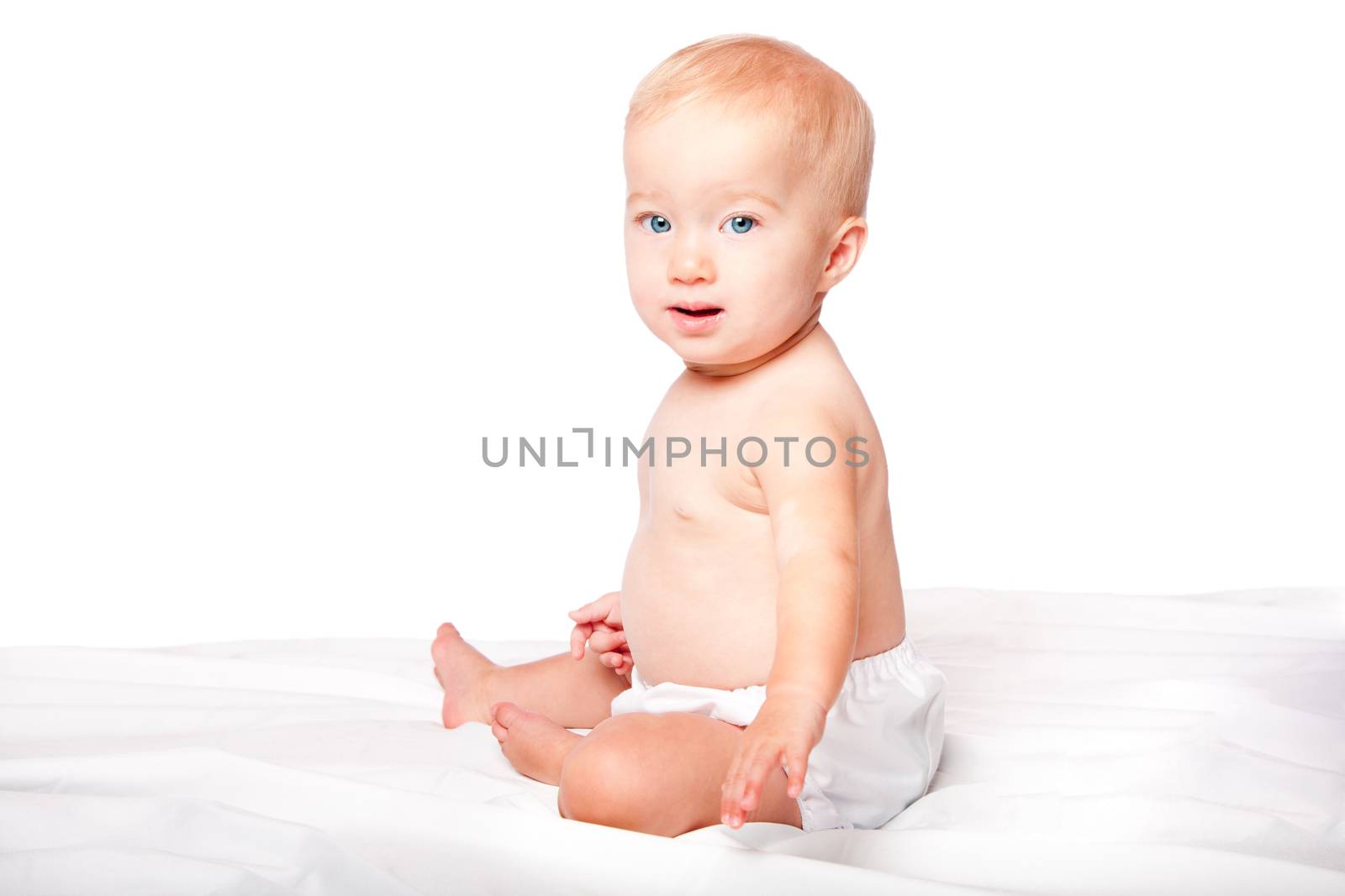 Cute blond baby with blue yes sitting on soft bed wearing diapers, on white.