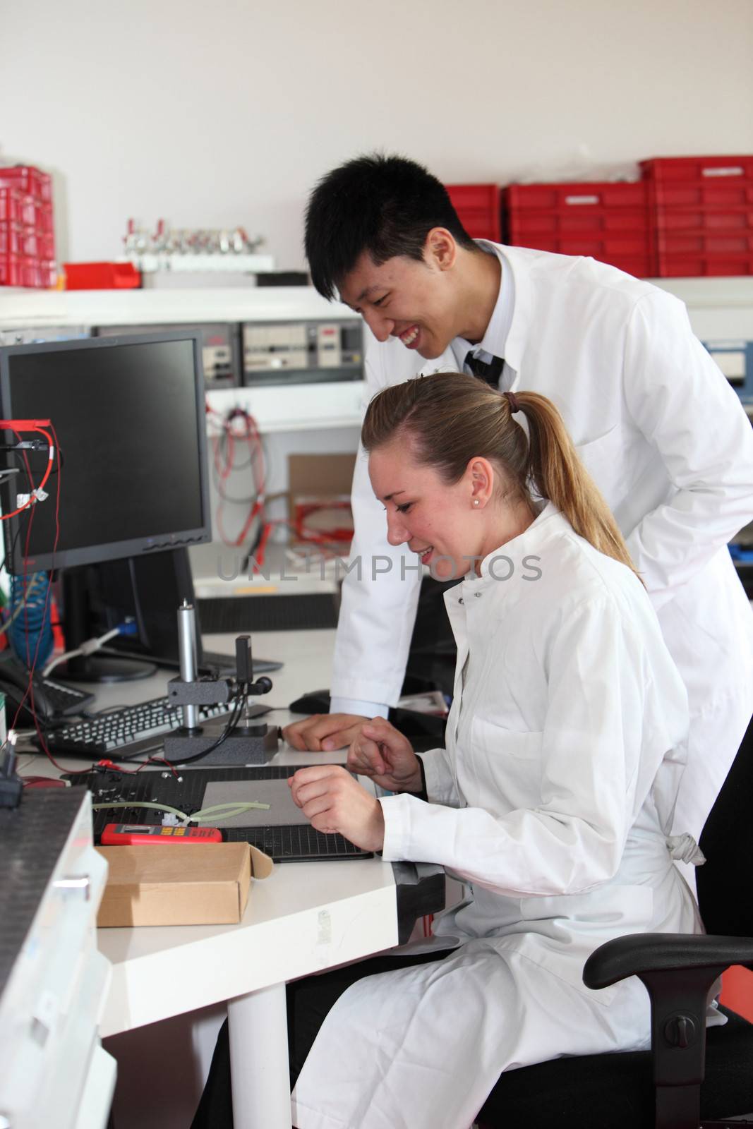 Two laboratory technologists at work, a young Asian man and his female colleague, standing over a piece of equipment waiting for a test to complete