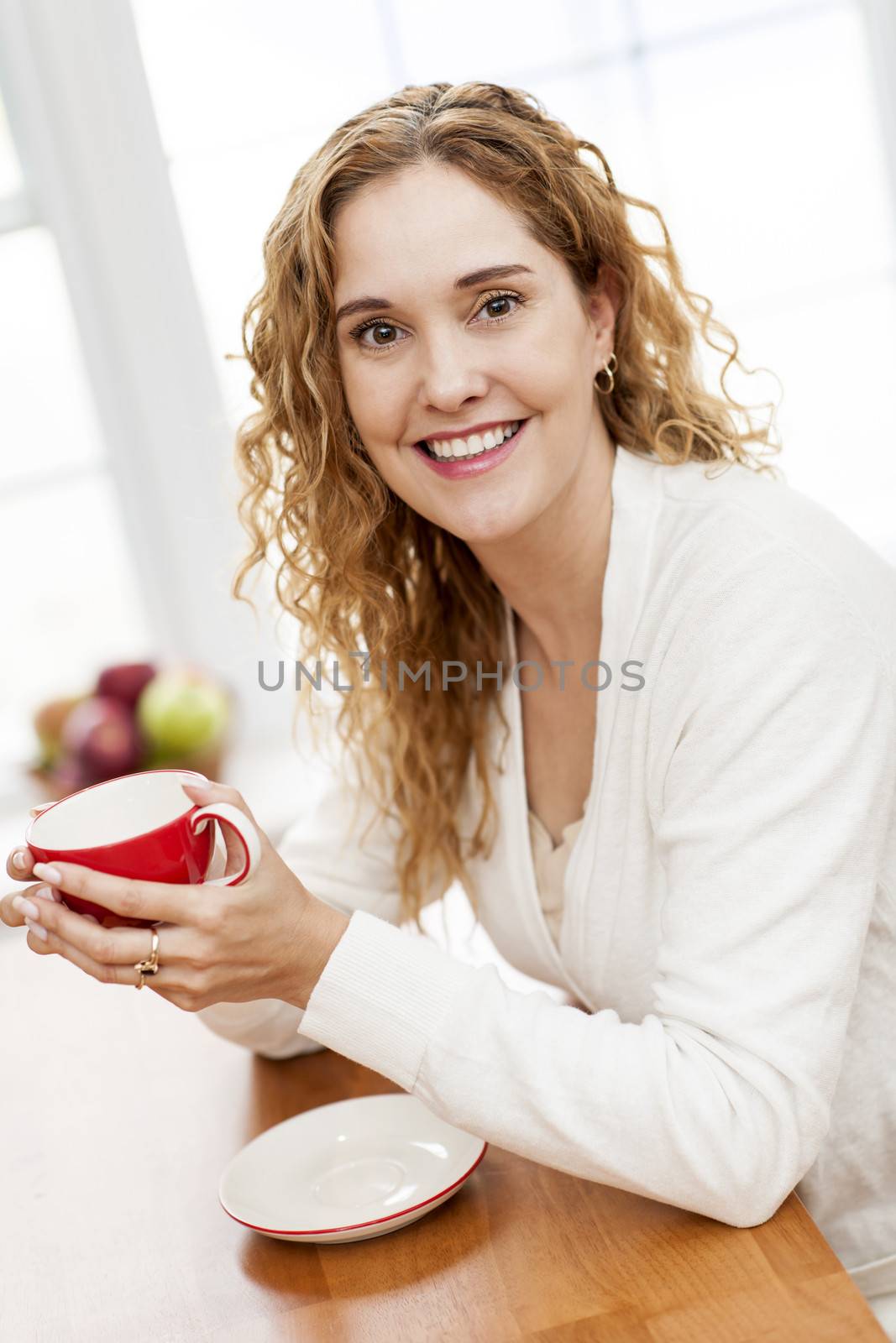 Portrait of smiling woman holding red coffee cup sitting at table in home kitchen by window