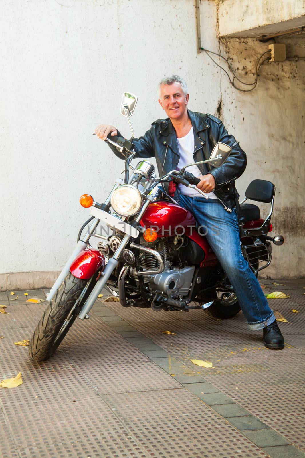 Vertical take of a smiling senior Caucasian male motorbike rider sat on a red cruiser wearing leather jacket in a relaxed pose. Generic shot location Bombay India