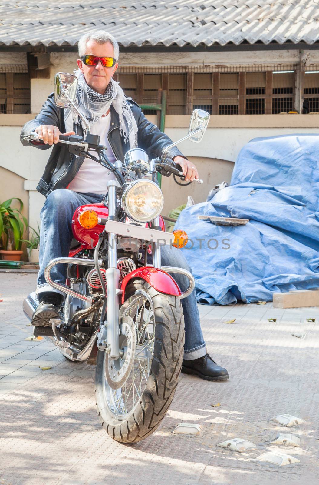 Color vertical portrait of a trendy fashion driven senior motor cyclist sat back on  a red cruiser smiling neutral expression in leather, denim, scarf and sunglasses. Generic shot location Bombay India with model release