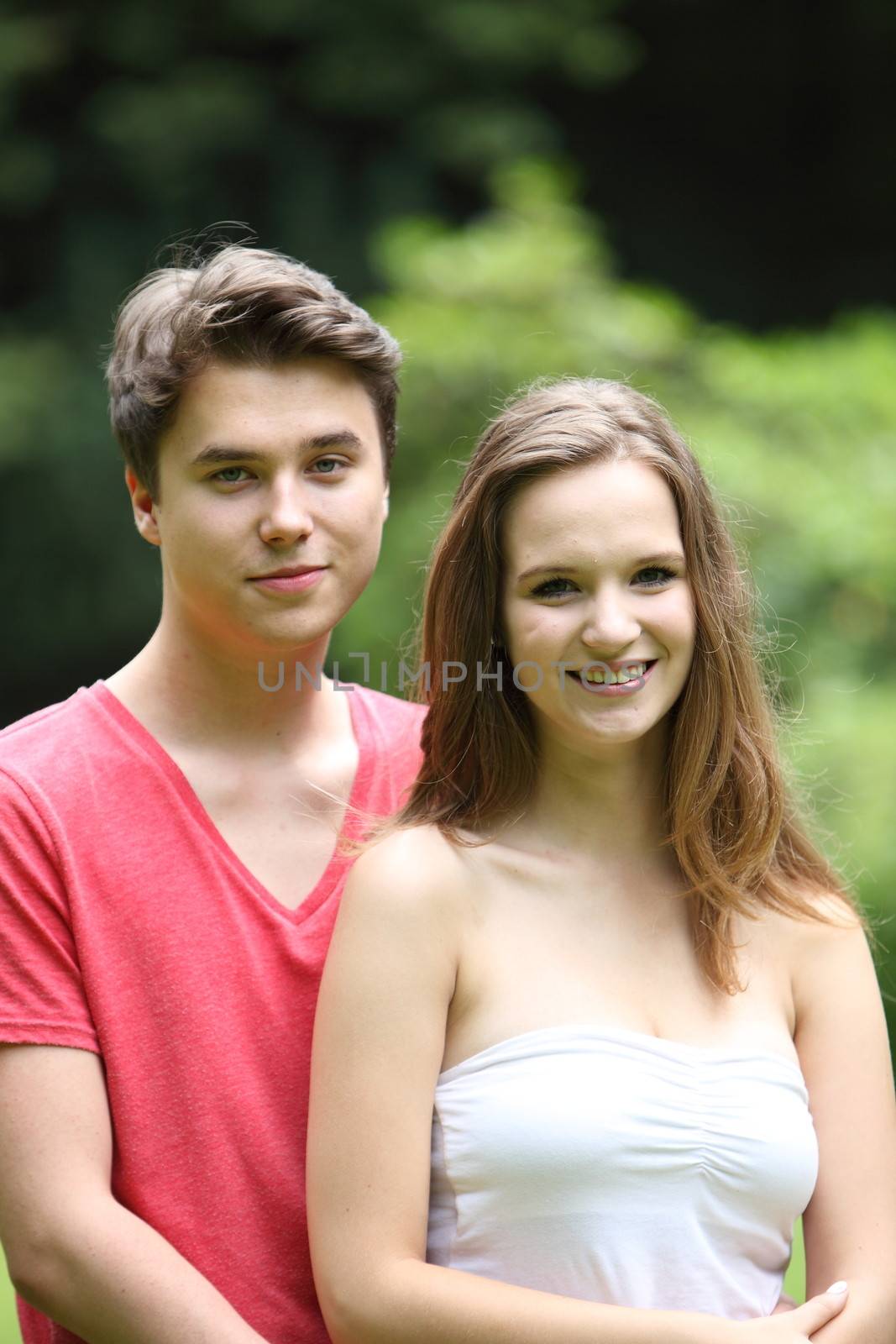 Beautiful young teenage couple standing close together smiling happily at the camera outdoors in a green park in the sunshine