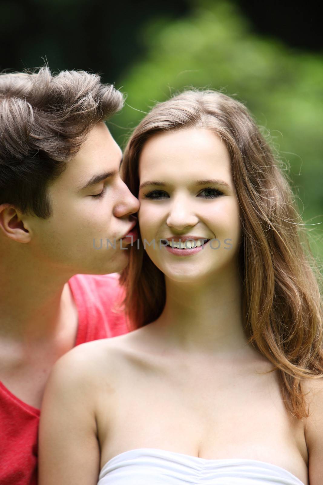 Teenage boy kissing his pretty young girlfriend on the cheek in a tender moment, close up portrait outdoors