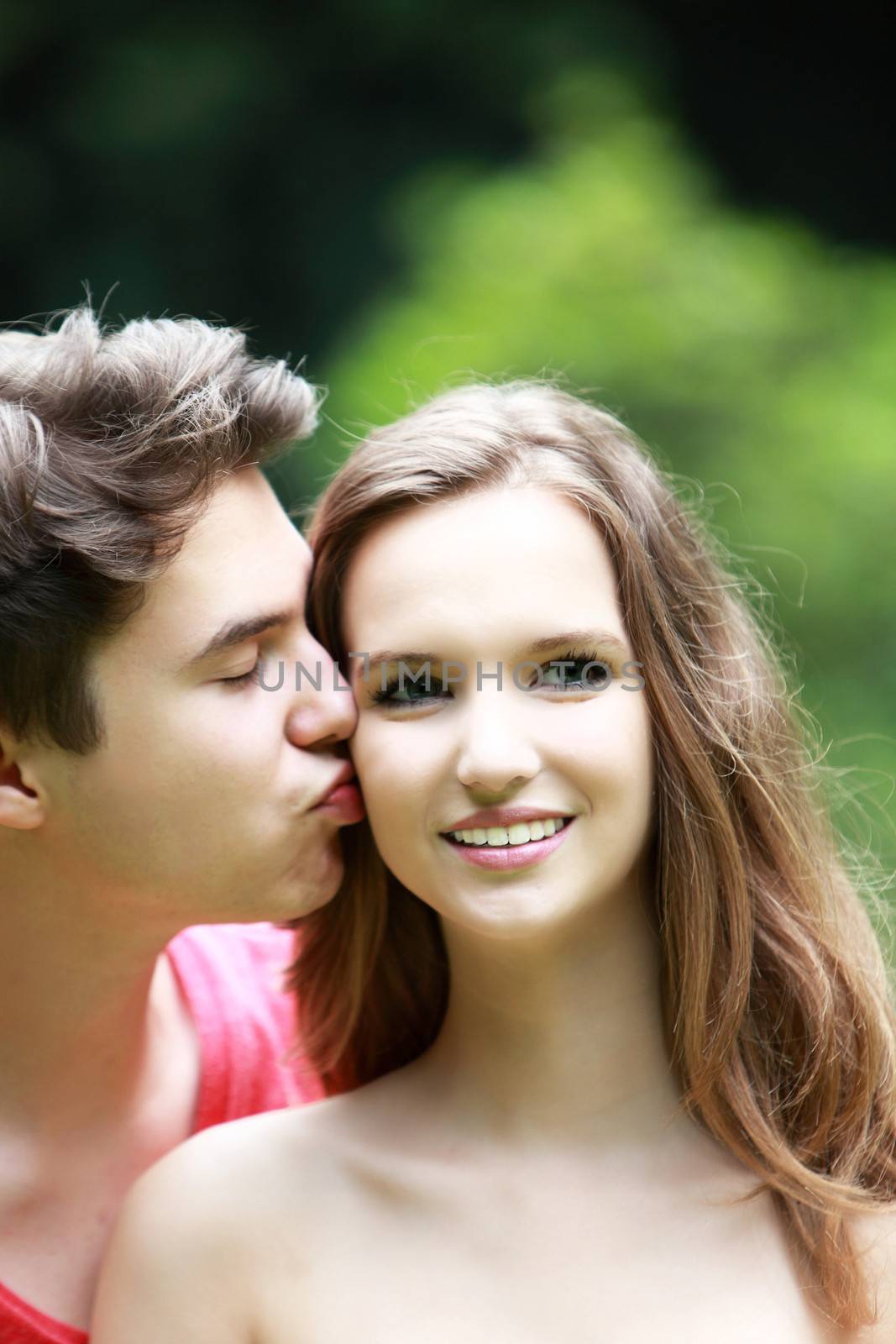 Handsome young man kissing his pretty teenage girlfriend outdoors against greenery