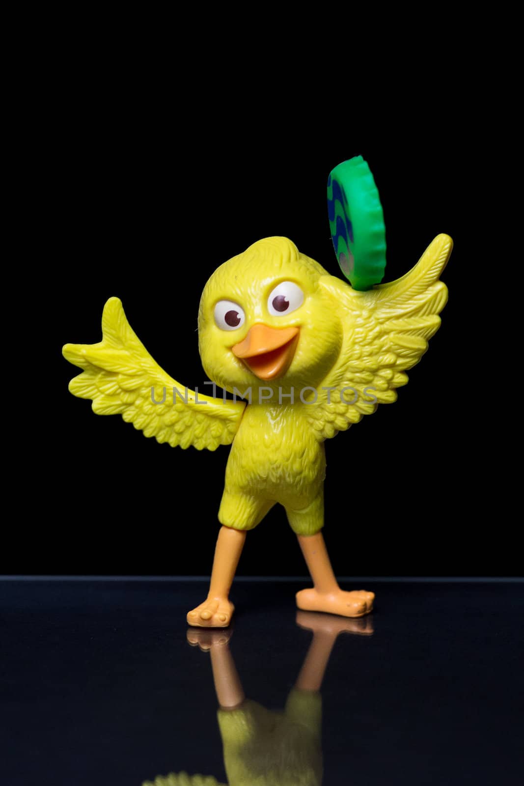A Yellow toy bird with a happy face holding a green object on a reflective black surface with black background