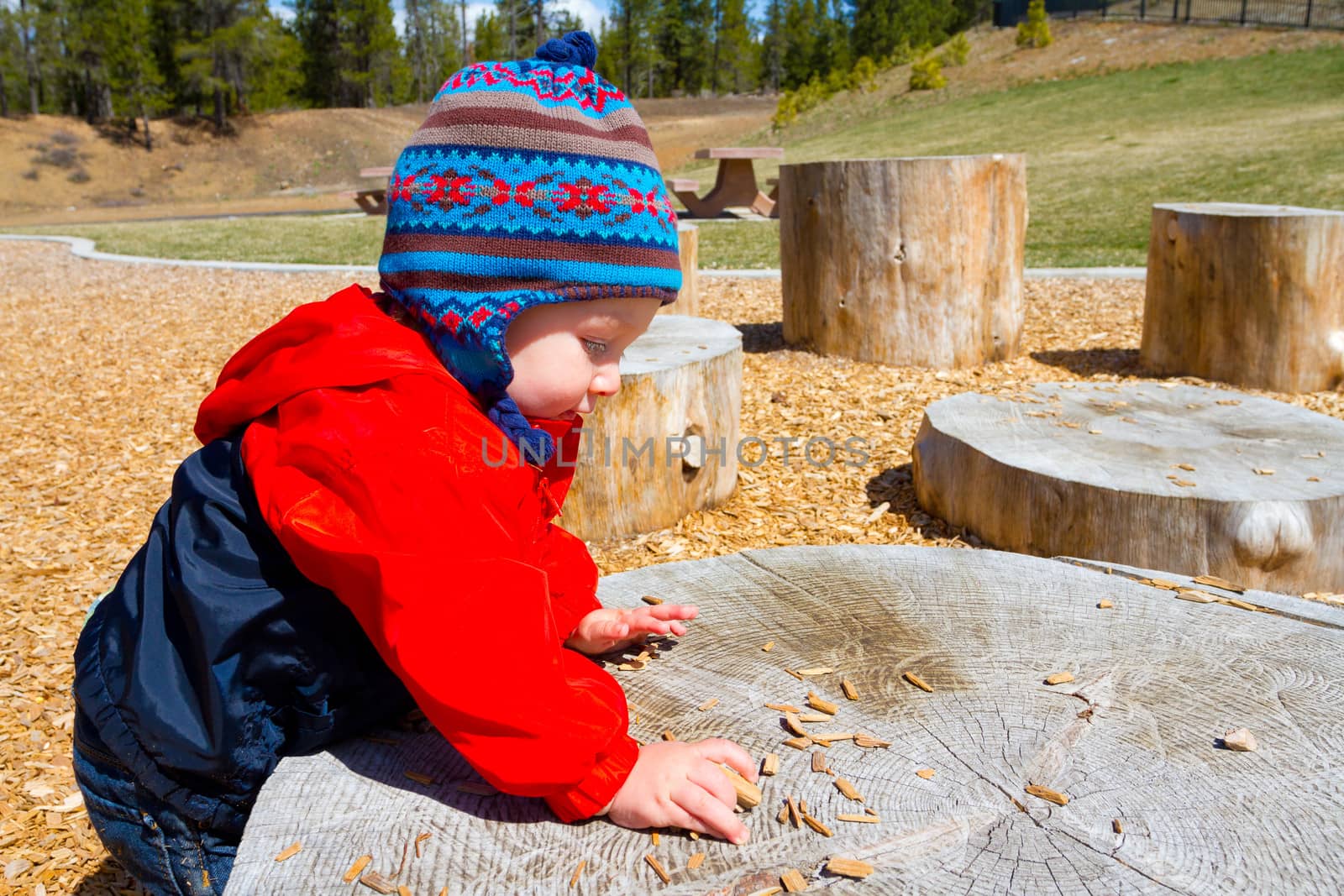 A one-year-old boy plays at a park outdoors with wood chips and structures to keep him entertained.