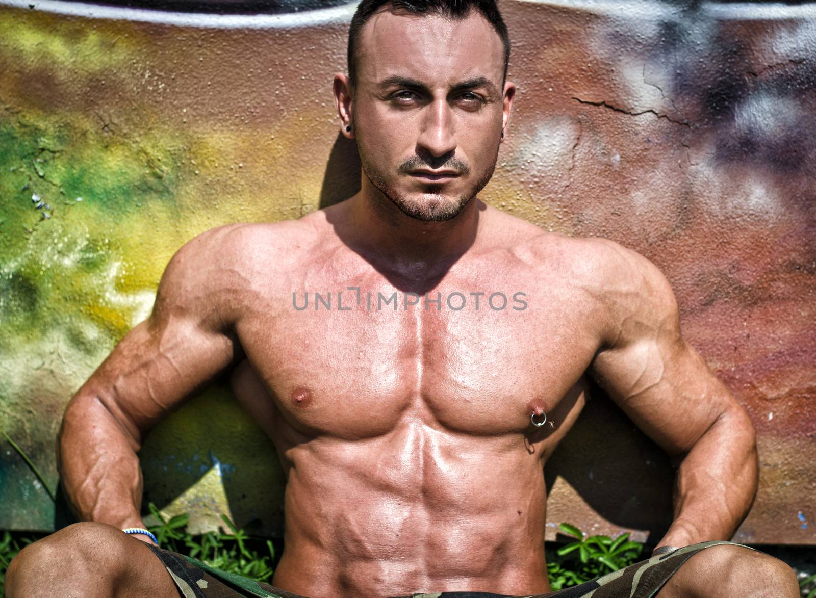 Muscular bodybuilder sitting against colorful wall showing ripped pecs and abs