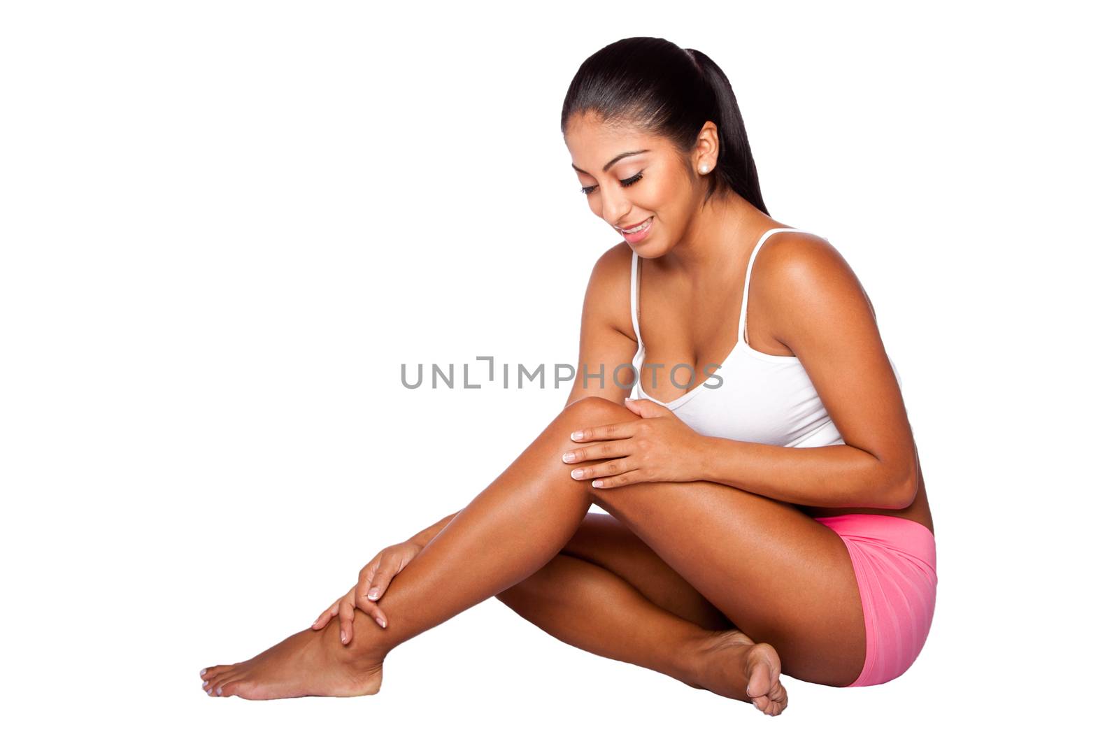 Beautiful happy smiling healthy young woman with radiant skin sitting on floor wearing sporty clothes holding caressing leg, bodycare concept.