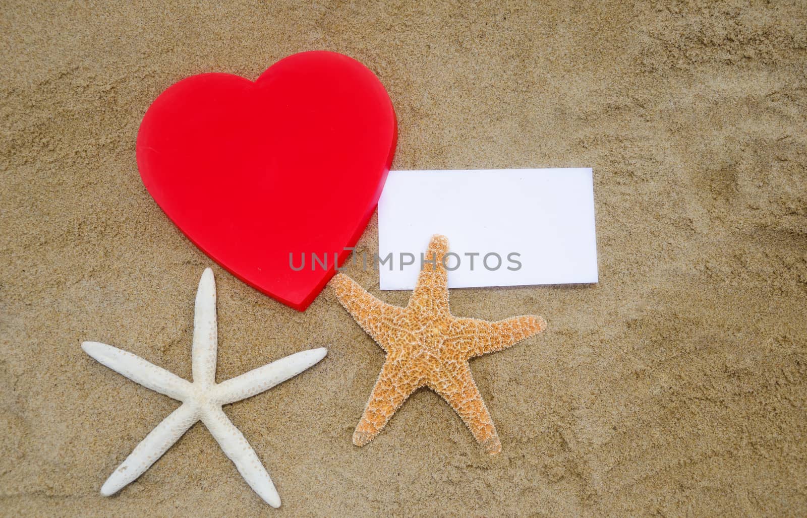 Red Heart shape, two starfishes, and piece of paper on the sandy beach