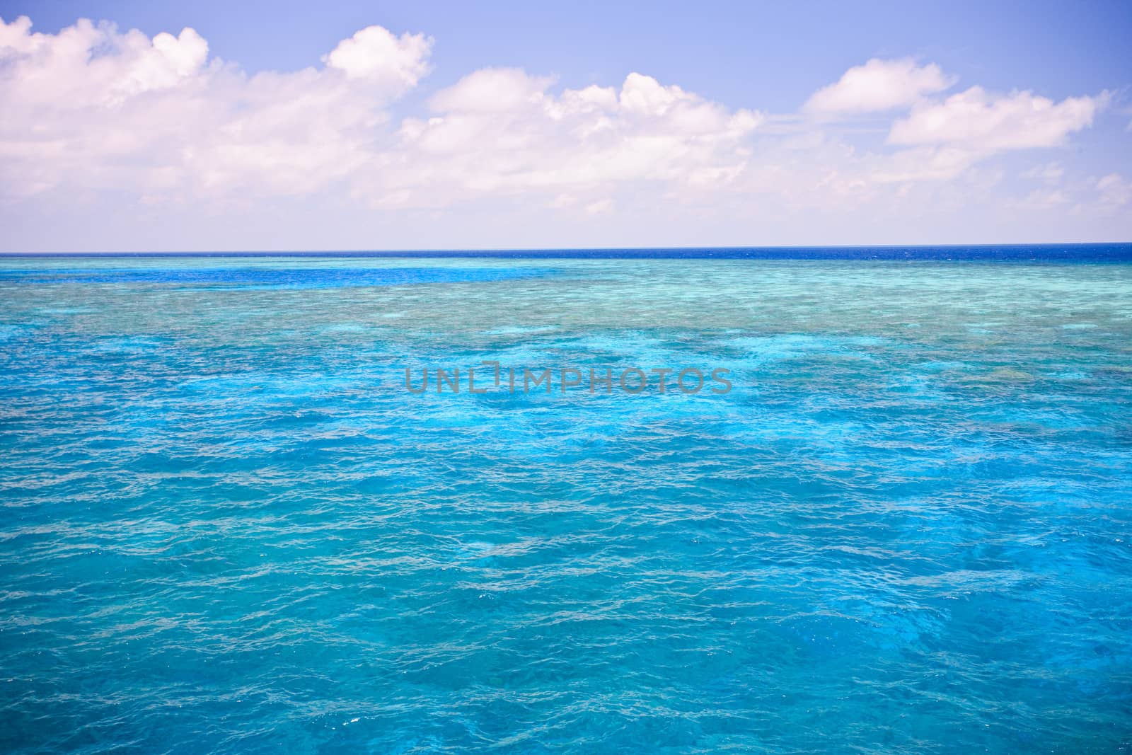 Background image of an azure blue ocean with shallow submerged offshore reef under a cloudy blue summer sky