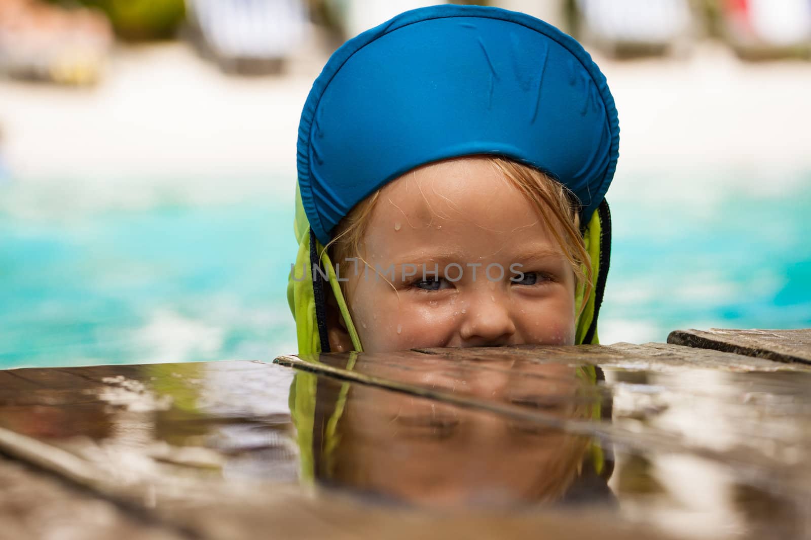 A cute young boy playing in water and peeking over side of pool.