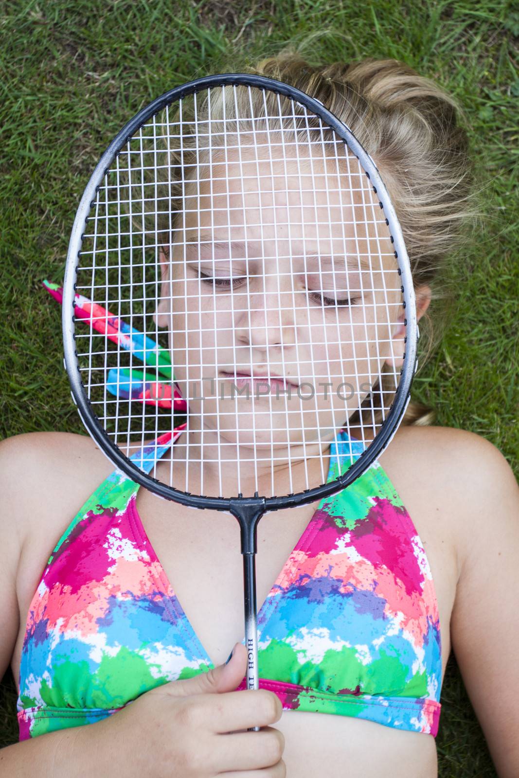 A teenage girl lying on the lawn outside with eyes closed, holding a badminton racket in front of her face