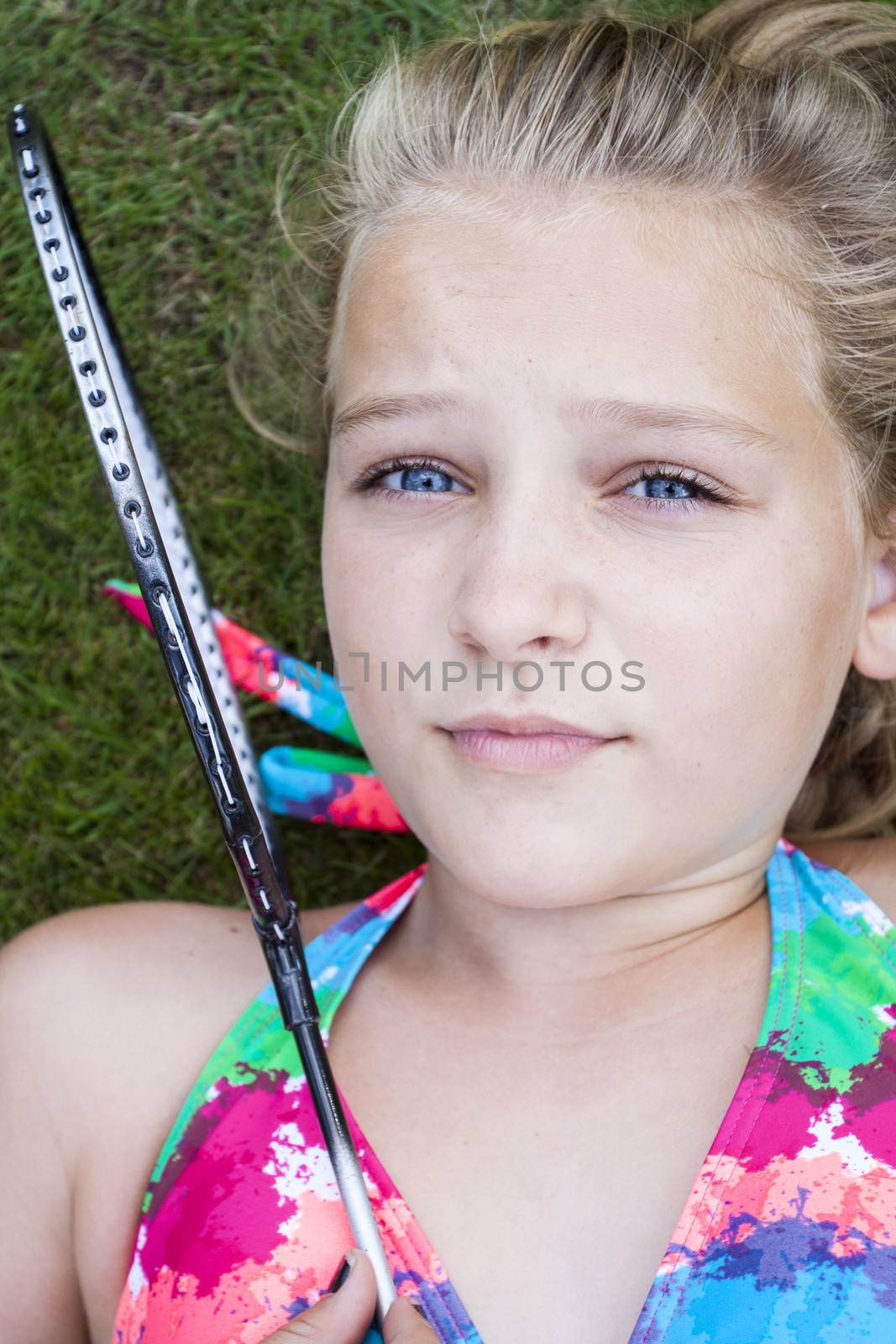A teenage girl lying on the lawn outside looking at camera, holding a badminton racket in front of her face