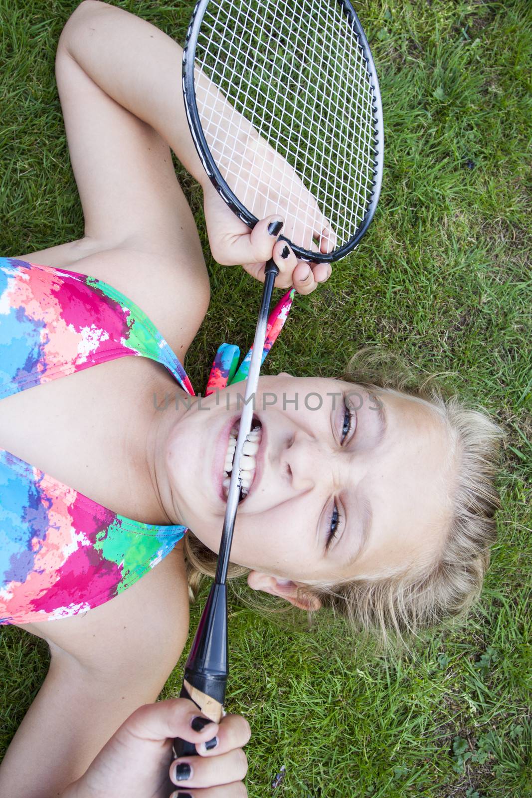 A teenage girl lying on the lawn outside, biting a badminton racket in frustration, doing a funny expression