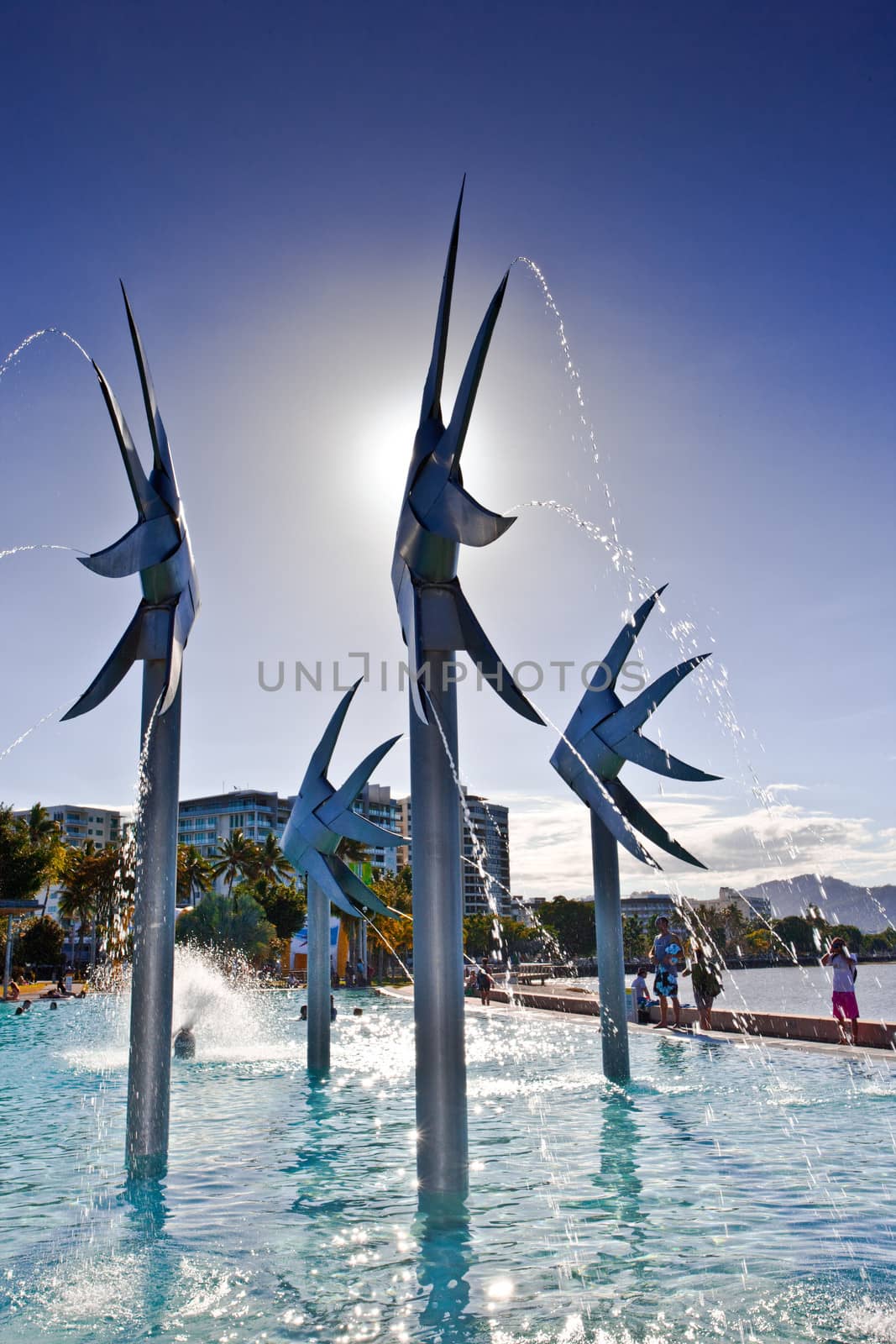 Steel fish sculpture and fountain at the Esplanade in Cairns with people playing in the water of the swimming lagoon on a hot summer day