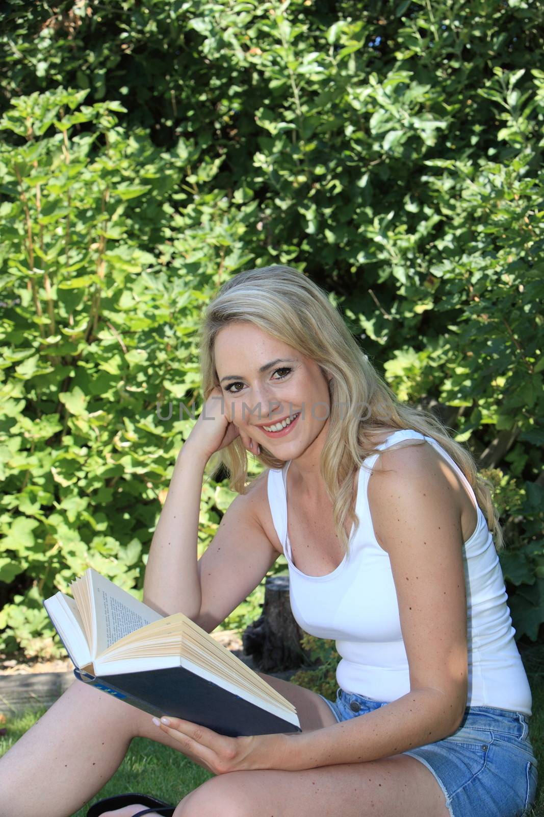 Pretty smiling young blond woman wearing summer shorts reading in the garden in the shade of a leafy green tree