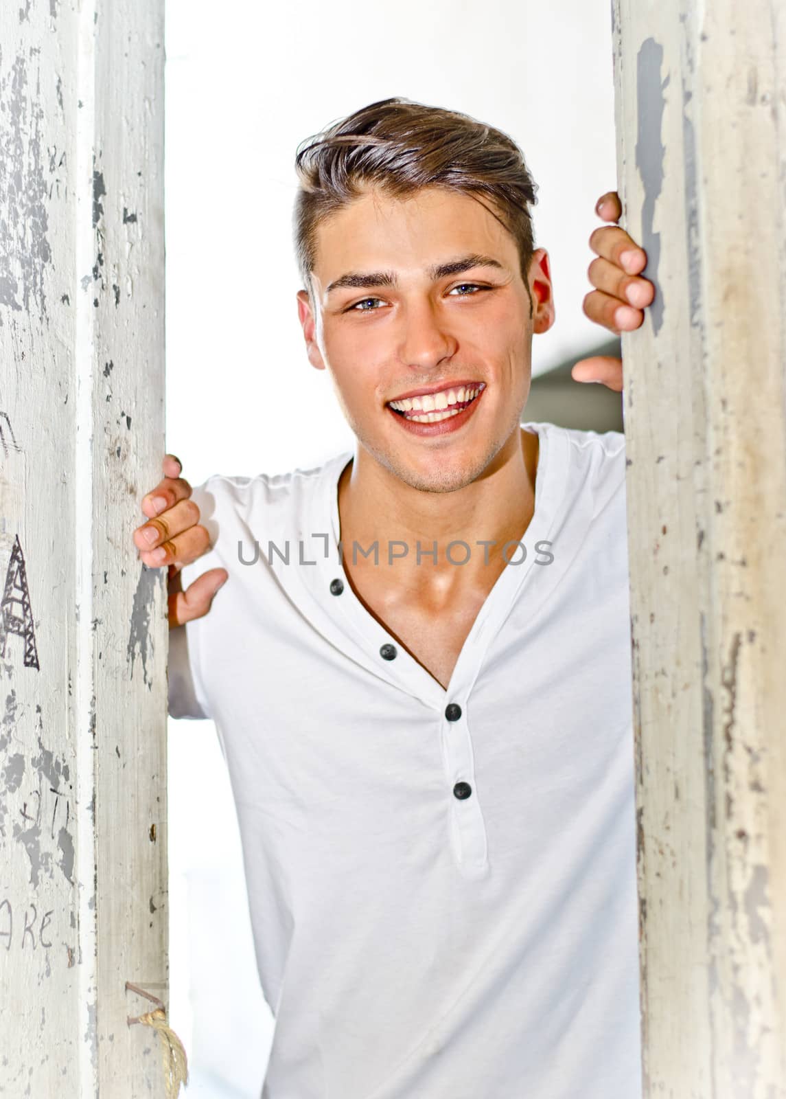 Happy, smiling young man in white between wooden doors looking in camera