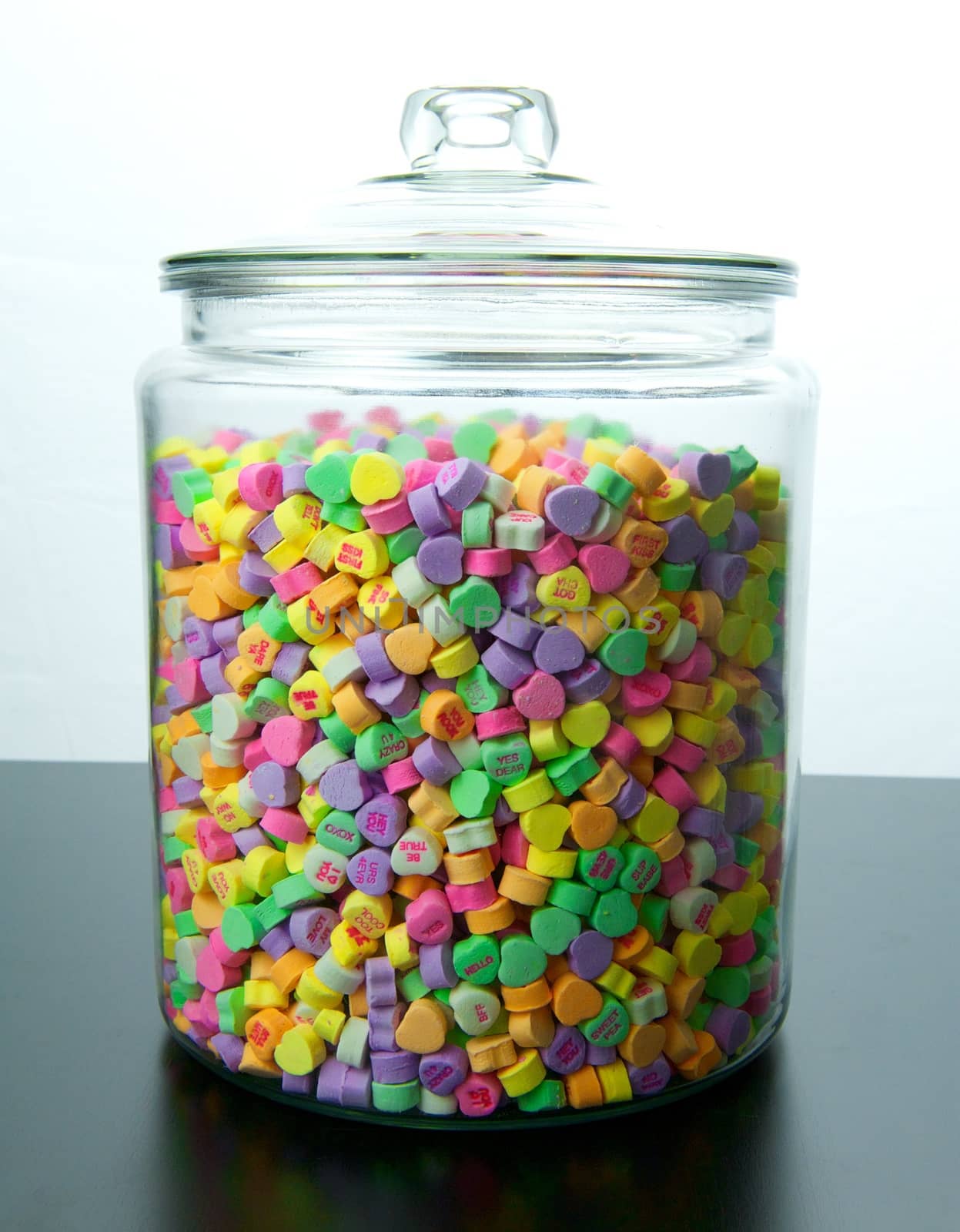 Large glass jar of colorful candy hearts with lid sits on a black table with a white background