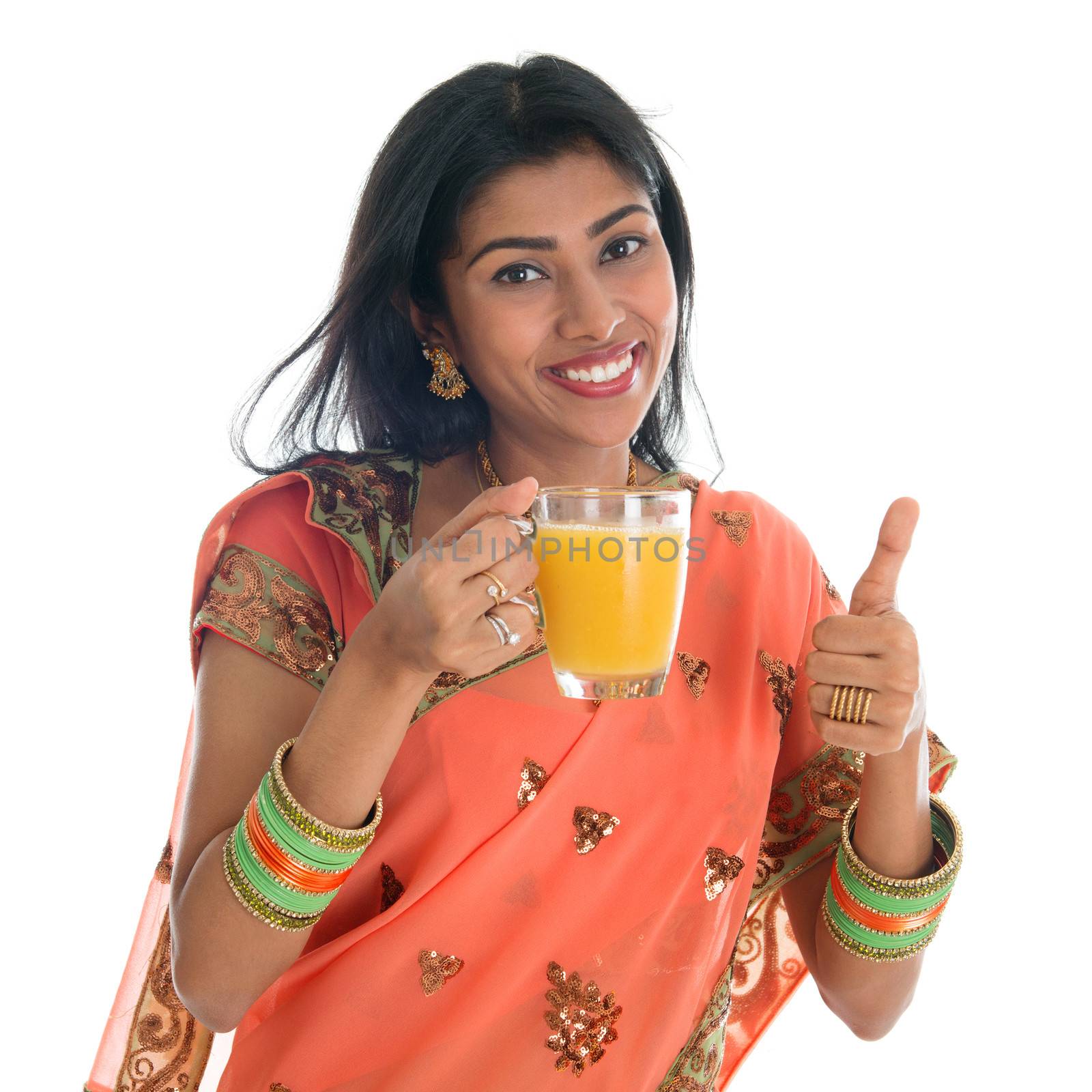 Happy traditional Indian woman in sari drinking a glass of orange juice showing thumb up, isolated on white background.