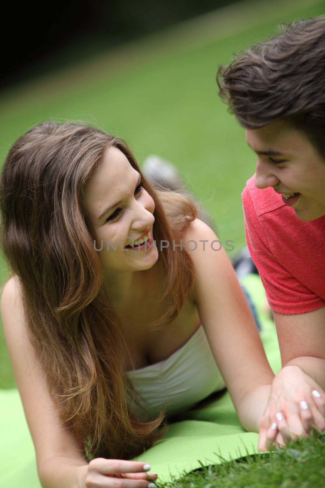 Young teenage girl on a date lying on the grass in a park holding her boyfriends hand smiling and chatting